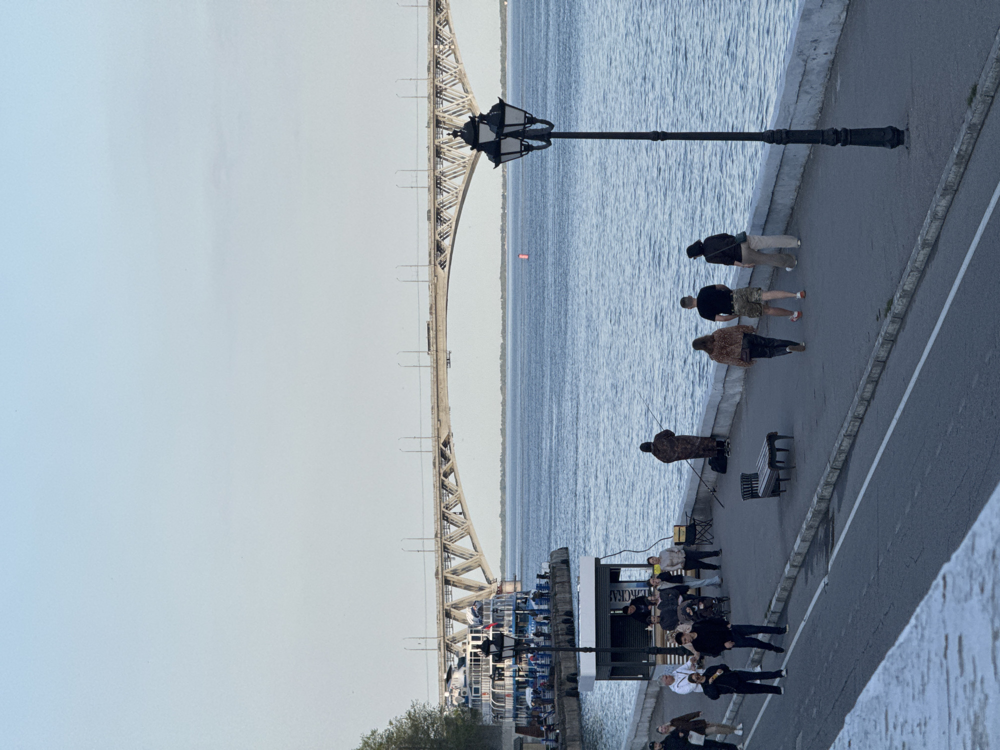The image depicts a waterfront scene in Saratov, Russia, with the Saratov Bridge spanning the Volga River in the background. The view is from an elevated position, possibly a quay or embankment. The bridge is a large, steel structure with a distinctive arched design. The river is visible to the side, with light ripples.
Along the quay, people are walking and congregating. Some individuals are strolling along the paved walkway, while others appear to be fishing near the water's edge. Several benches are present along the walkway. A small building stands near a docked passenger ferry. A vintage streetlight with ornate glass lanterns is placed between the pavement and the river.