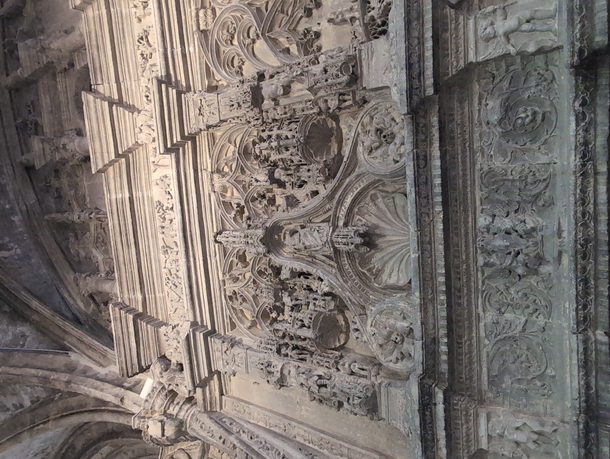 The image depicts a section of the intricately carved facade of the Collegiate Church of Saint-Jacques in Le Tréport, France. The stone facade is highly detailed with Gothic architectural elements and sculptural ornamentation. The top portion of the image shows the ribbed ceiling of the church, while the main focus is on a large, ornate stone relief. This relief features multiple tiers of detailed carvings, including figures, architectural motifs, and possibly biblical scenes. The central area has a prominent shell-like design, framed by elaborate tracery and smaller figures. Below this, a frieze runs across the facade, displaying more relief work with floral and figural elements, including what appear to be cherubic figures on the sides. The stonework is weathered and aged, suggesting it is an ancient structure. The lighting is dim, casting shadows that accentuate the depth and texture of the carvings, creating a somber and impressive atmosphere.