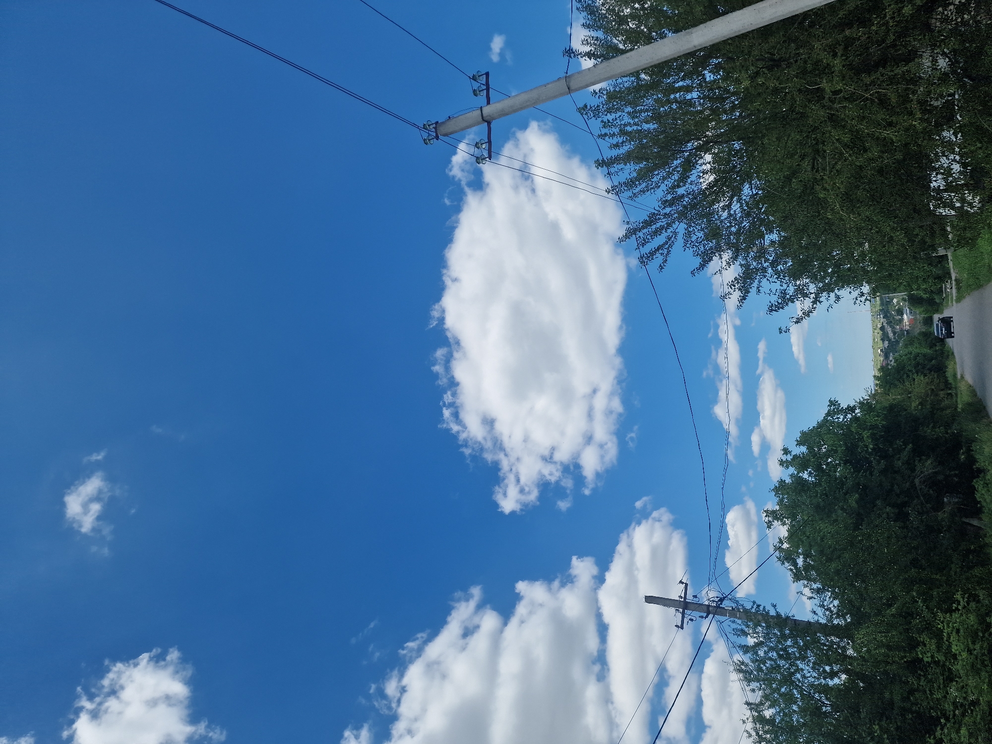 The image captures a rural road scene under a bright blue sky with scattered white clouds. Two utility poles with visible wires are present, one on the left and another more prominently on the right. Dense green trees line both sides of the road, creating a natural canopy. The road itself is paved and leads into the distance where a small dark-colored car is visible. In the background, a small settlement with houses and some vegetation is discernible. The overall impression is one of a tranquil, sunny day in a countryside setting.