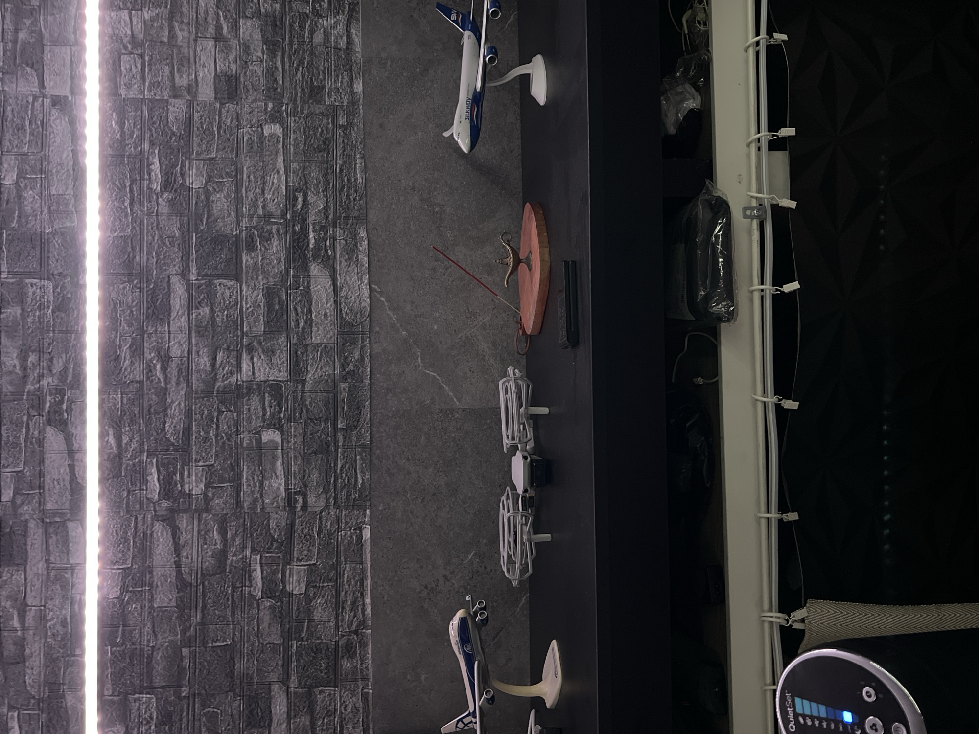 This is a forensic, matter-of-fact description of a scene, located in Schoonderbuken, Belgium.
The image depicts a shelf against a dark, textured wall, possibly wallpaper designed to resemble brick. A strip of LED lights illuminates the top portion of the wall. On the shelf, several objects are arranged. To the left, a model airplane with white wings and a blue fuselage is displayed on a white stand. Next to it, a small white drone with protective cages around its propellers is positioned. Further to the right, a small, ornate brass lamp, resembling a genie lamp, sits on a red, heart-shaped wooden base. An incense stick is inserted into the lamp, with smoke rising from its tip. To the far right, another model airplane, similar in design to the first but with "SILKWAY" written on its fuselage, is also on a white stand. Below the shelf, the image shows the top of a white unit with various wires and clips attached to it, and a black patterned surface below that. On the far left, a circular device with a digital display and buttons is visible, labeled "QuietSet". The overall environment suggests an indoor setting, possibly a display or storage area.