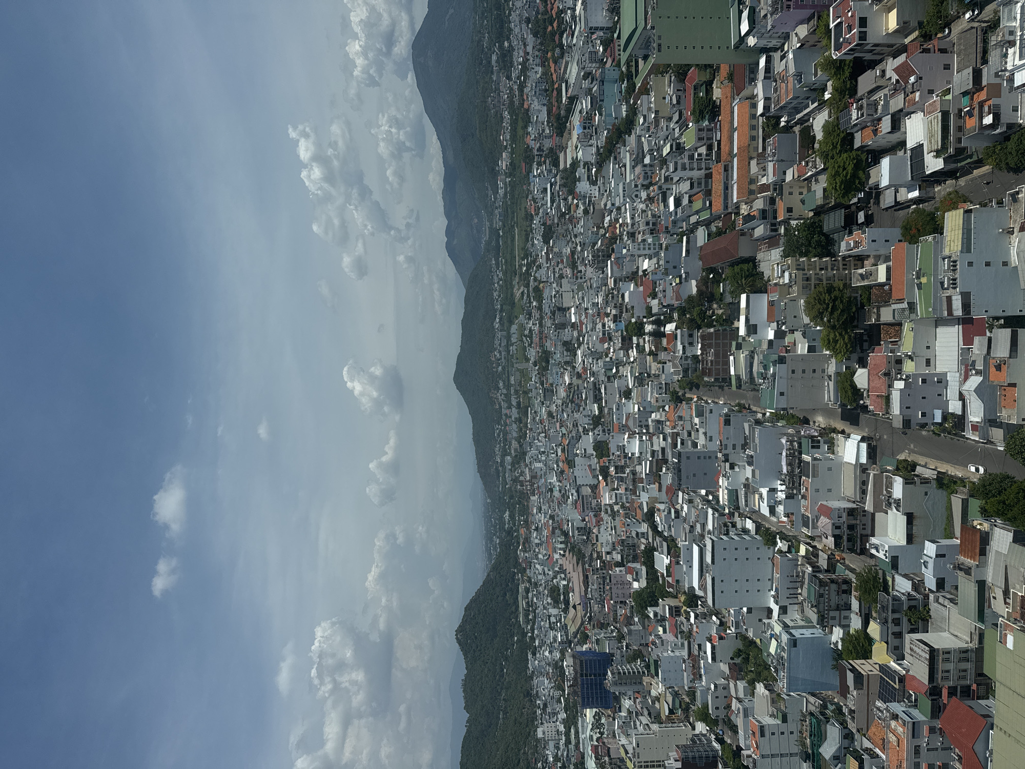 An aerial view captures a dense urban landscape with a backdrop of lush green mountains under a partly cloudy sky. The city, identified as Bac Nha Trang Ward, Vietnam, is characterized by a tightly packed arrangement of buildings, predominantly low-rise structures with a mix of residential and commercial properties. Rooftops display a variety of colors, including white, grey, orange, and green, with some featuring solar panels. Streets wind through the city, with a few vehicles, including a white car and a motorcycle, visible. The architecture is diverse, with modern multi-story buildings interspersed with older, more traditional homes. The mountainous terrain in the distance is covered in dense vegetation, creating a stark contrast with the urban sprawl. The sky is a bright blue, with scattered white cumulus clouds adding depth and texture to the scene. The overall impression is one of a vibrant, growing city nestled within a scenic natural environment.
