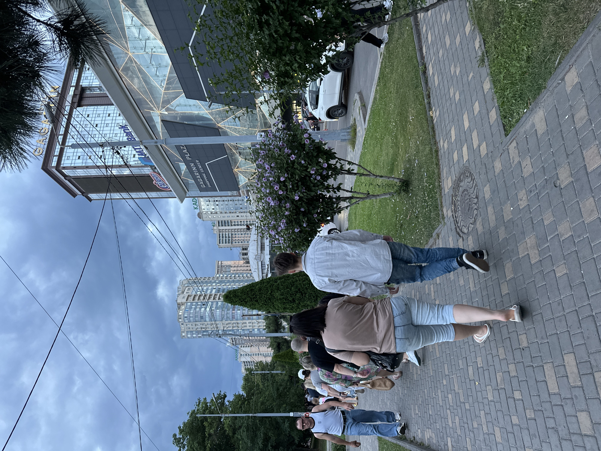 The image captures a scene on a sidewalk in front of the Gagarin Plaza shopping center in Odesa, Ukraine. A group of people, seen from the back and side, are walking along a paved sidewalk. The people are casually dressed, with some wearing jeans and t-shirts, and others in lighter clothing. A white car is visible on the street to the right, and green trees and bushes line the sidewalk and the grassy area next to it. The Gagarin Plaza shopping center, a modern building with glass facades and signage including "GAGARINN PLAZA SHOPPING CENTER" and "MT Bank", is prominent in the background, along with several tall apartment buildings. The sky is overcast with dark clouds, suggesting an impending storm or a late afternoon light.