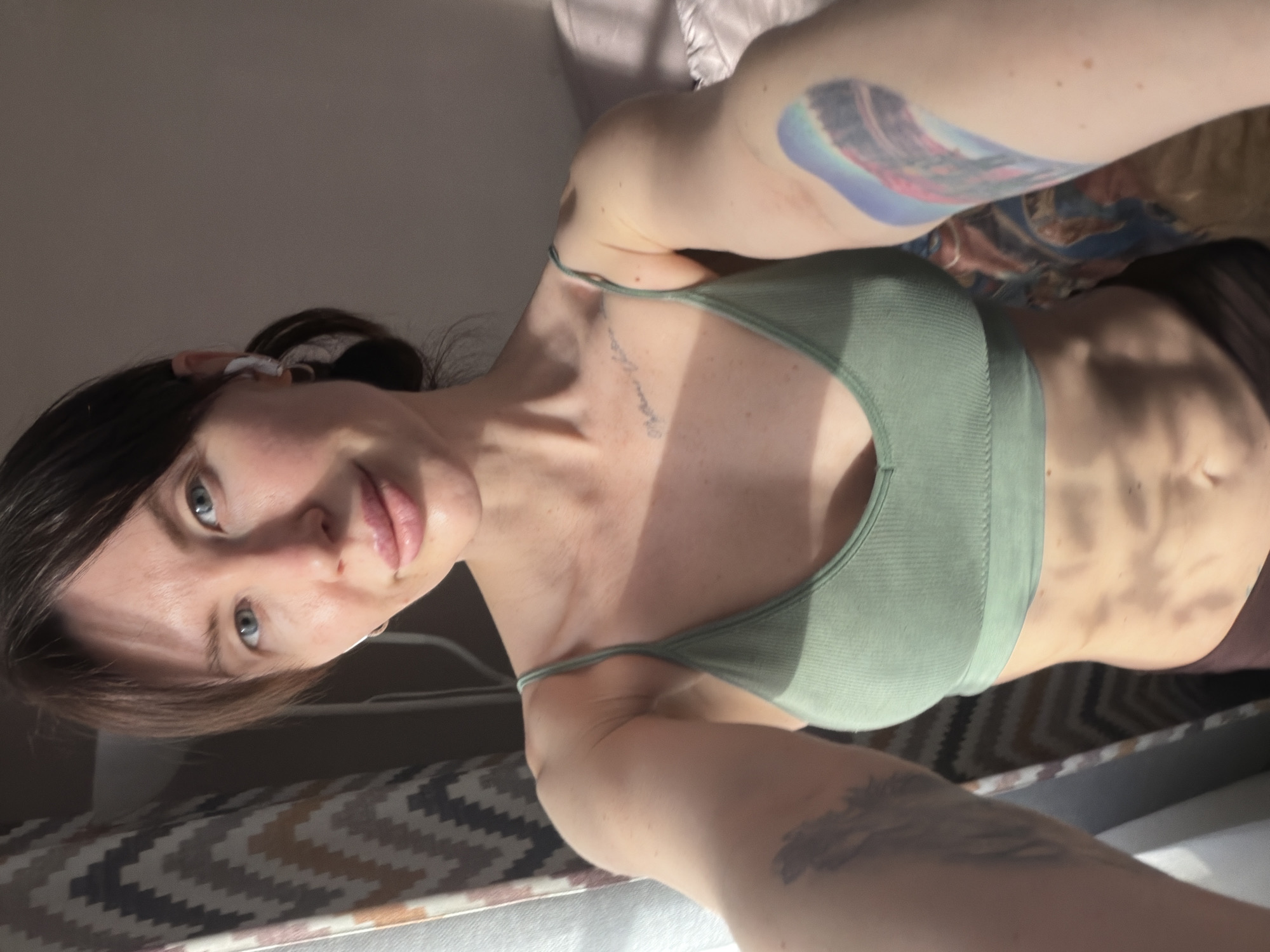 A head-on, slightly high-angle selfie captures a woman with dark hair pulled back, fair complexion, and blue-grey eyes, looking directly at the camera with a neutral expression. She is wearing a light green ribbed sports bra or crop top. Several tattoos are visible: a colorful, possibly abstract landscape design on her upper left arm, a dark, script-like tattoo on her left collarbone/chest, and an indistinct dark tattoo on her right forearm. A small earbud is present in her left ear.
The setting is indoors, likely a room bathed in natural sunlight, casting strong, distinct shadows across her torso, highlighting her abdominal muscles, and onto the light-colored walls in the background. In the lower foreground, a patterned textile with a geometric chevron design (brown, beige, and black) is visible, possibly a blanket or rug. Soft, light-colored bedding or fabric is seen behind her head and shoulders. The strong shadows suggest it is daytime, possibly morning or late afternoon. No legible text is visible in the image. The general location context is Perm, Russia.