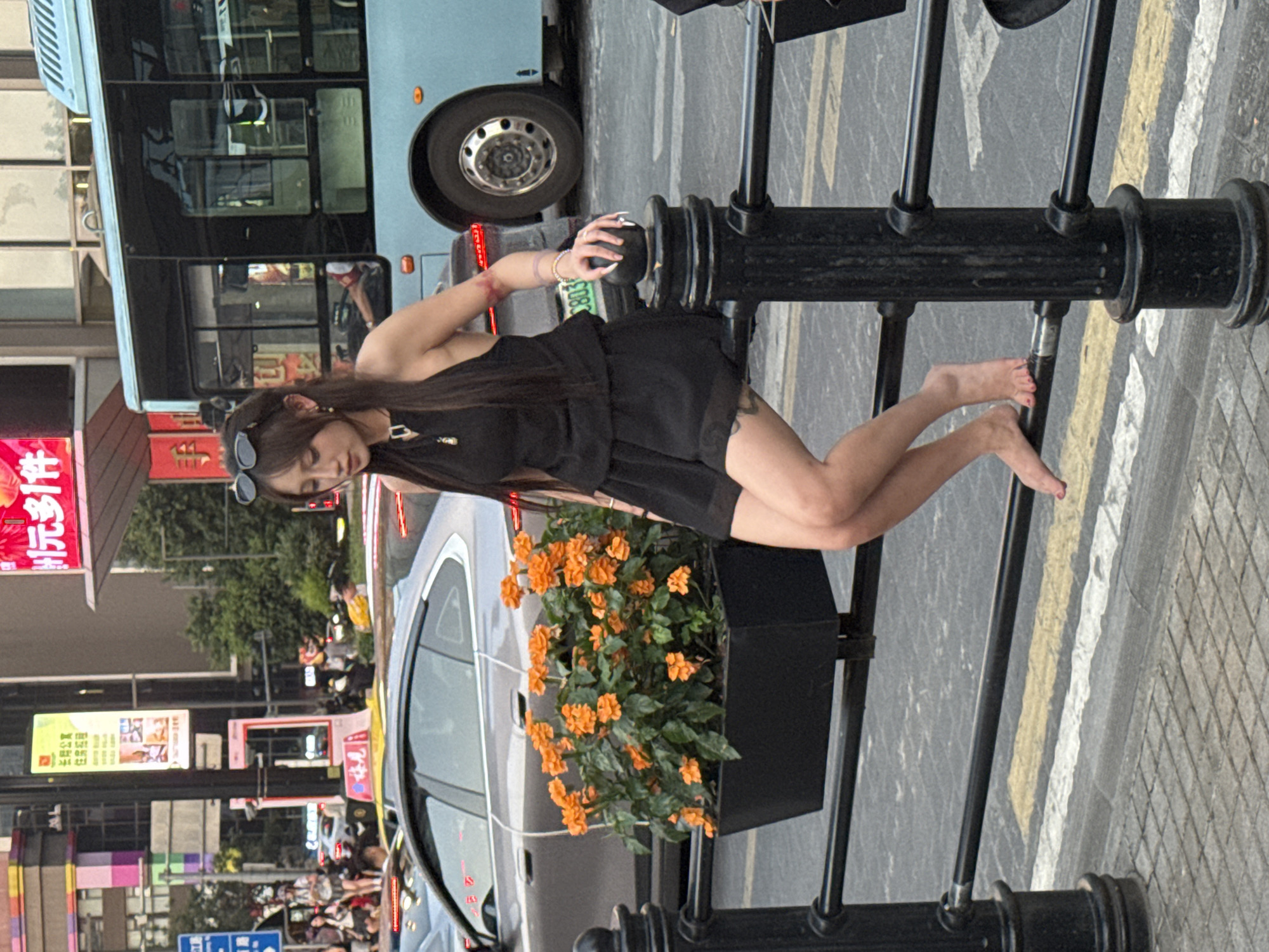 A young woman with long dark hair and sunglasses perched on her head is sitting on a black metal railing in the Yuzhong District of Chongqing, China. She is wearing a black halter top and black shorts, and her bare feet are dangling below the railing. She has long fingernails, some of which are painted. She is looking down and to her right. Behind her, a light blue bus is visible, along with a silver car. In front of the railing, a planter box filled with vibrant orange flowers is placed. The scene appears to be a street in a busy urban environment, with buildings and traffic visible in the background.