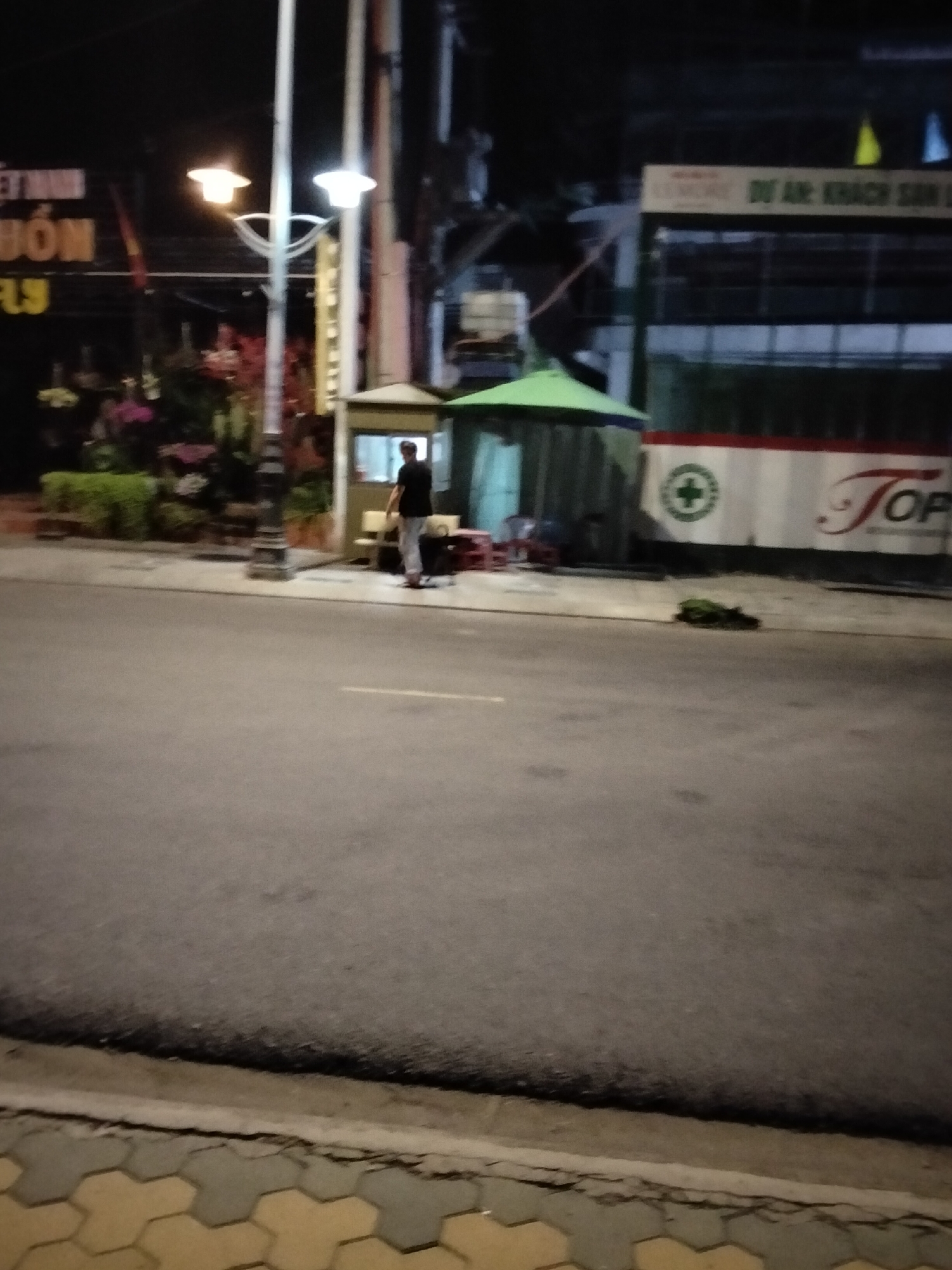 The image shows a street scene at night in Mũi Né, Vietnam. In the foreground, a paved road and sidewalk are visible. Further back, a single individual, appearing to be a man wearing a dark shirt and light-colored pants, stands near a small booth or kiosk with a green awning. Behind him, a building with signage that includes "DU AN KHACH SAN" and a logo with a green cross and the word "TOP" is visible. The scene is illuminated by streetlights and lights from the buildings. Vegetation and flowers are present to the left of the kiosk. The overall impression is of a quiet street in a Vietnamese town at night.