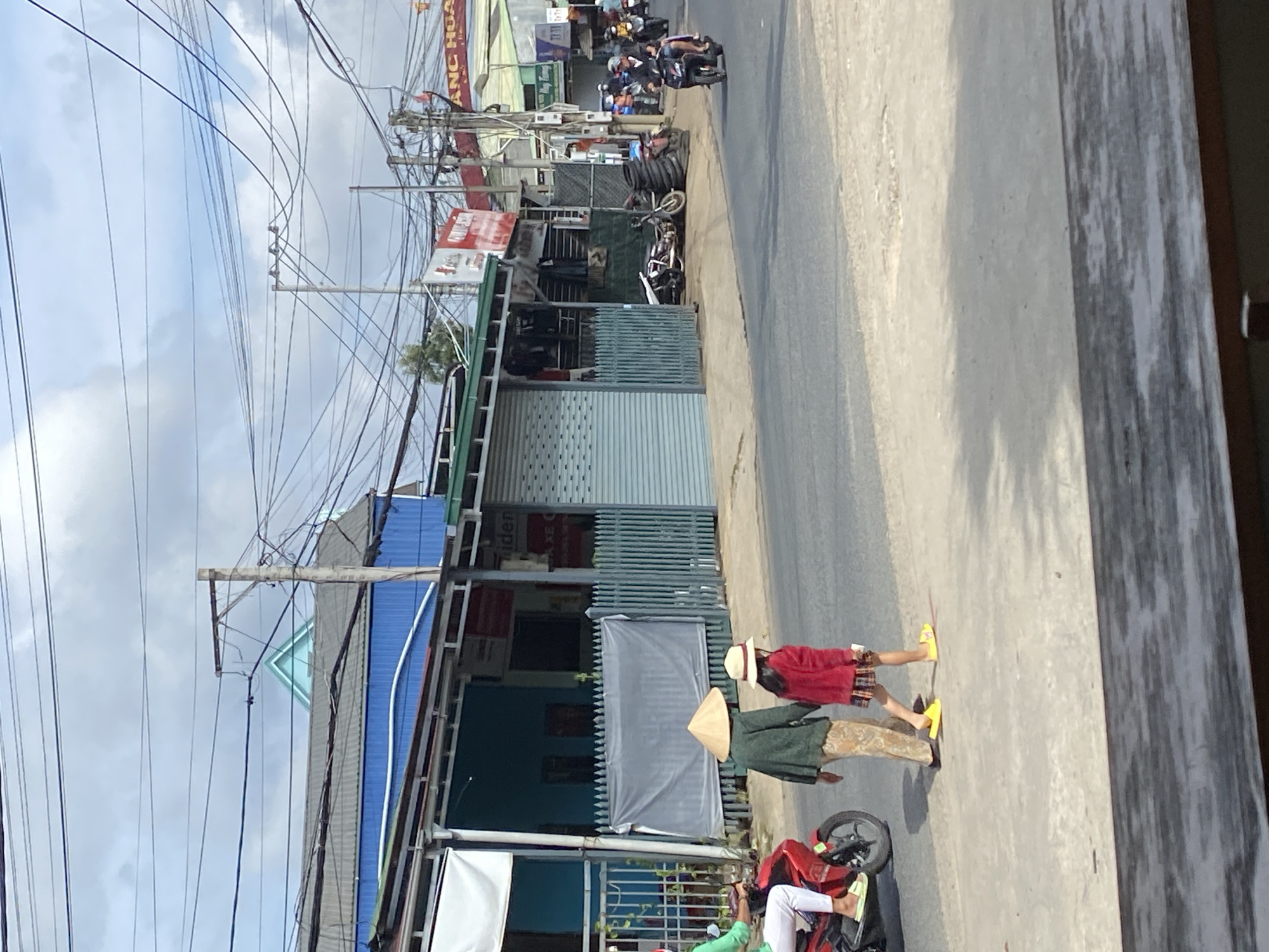 The image depicts a street scene in Da Lat, Vietnam, characterized by a mix of residential and commercial buildings lining the road. In the foreground, two individuals are walking on the sidewalk. One, likely an adult, wears a conical hat and a green garment over patterned pants. Beside them, a child, possibly a girl, wears a red jacket and a plaid skirt, also with a hat. They are walking away from the viewer, towards the right side of the frame.
To the left, a red motorcycle is parked, with a person on it wearing a green shirt and white pants, facing towards the left. The street itself is paved, with a slight unevenness in the surface. Buildings on the right side of the street have a blue facade and metal roll-up doors, suggesting commercial establishments such as shops or workshops. Overhead, a complex network of electrical wires crisscrosses the sky, typical of urban Vietnamese landscapes. Further down the street, several motorbikes are visible, some with riders, indicating ongoing traffic. The buildings are adorned with various signs, some in Vietnamese. The overall impression is of a bustling, everyday scene in a Vietnamese city.