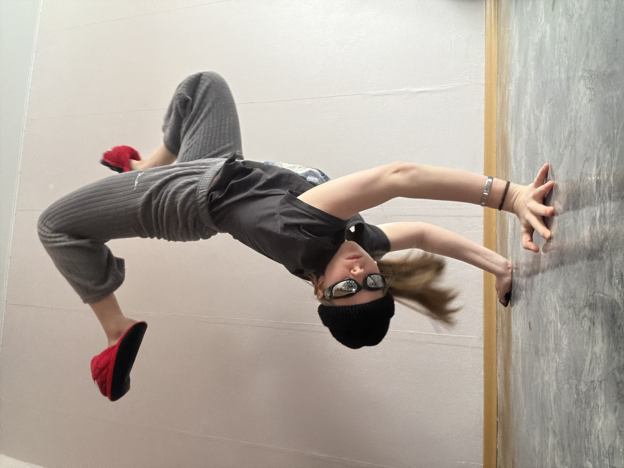 An indoor full-shot captures a young individual, likely a teenager or young adult, in an athletic or dance pose. The image is rotated 90 degrees clockwise, giving the impression that the person is suspended horizontally, parallel to the ground, with their head pointing downwards and feet elevated. If viewed correctly, the person is performing a backbend or bridge, arching their back while lying on a light-colored surface (likely the floor), with their hands pressed against a textured grey wall. Their head is positioned close to this textured wall, and their feet extend towards a plain, light grey/white wall.
The person is dressed in a black beanie, reflective sunglasses, a dark t-shirt, and loose-fitting grey ribbed pants, finished with bright red slippers. Accessories include a silver-toned bracelet on one wrist and a dark cord bracelet on the other. Their long, light brown hair flows downwards. The setting, located in Perm, Russia, is an interior space featuring two distinct wall textures: a plain light grey/white surface and a contrasting grey wall with a mottled, marble-like or concrete-like pattern, separated by a vertical yellowish trim. The lighting is soft and ambient, indicative of an indoor environment. The individual's facial expression, partially obscured by sunglasses, appears focused. There is no visible text in the image.