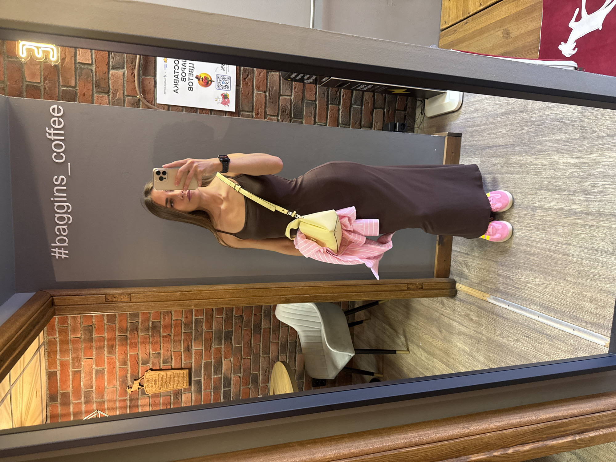 A young woman is taking a mirror selfie in what appears to be a coffee shop in Saint Petersburg, Russia. She is wearing a long, dark brown slip dress, bright pink sneakers, and carrying a small yellow handbag over her shoulder. A pink shirt is draped over her arm and handbag. The background features a brick wall, a gray wall with a neon sign that says "#baggins_coffee", and a wooden table with a chair reflected in the mirror. A wooden doorframe is visible to the left, framing a view into another part of the establishment. A decorative plaque with an image of an animal and text in Russian is mounted on the brick wall. A white, upright object, possibly a humidifier, is on the floor in the background. The floor is made of light-colored wood planks.