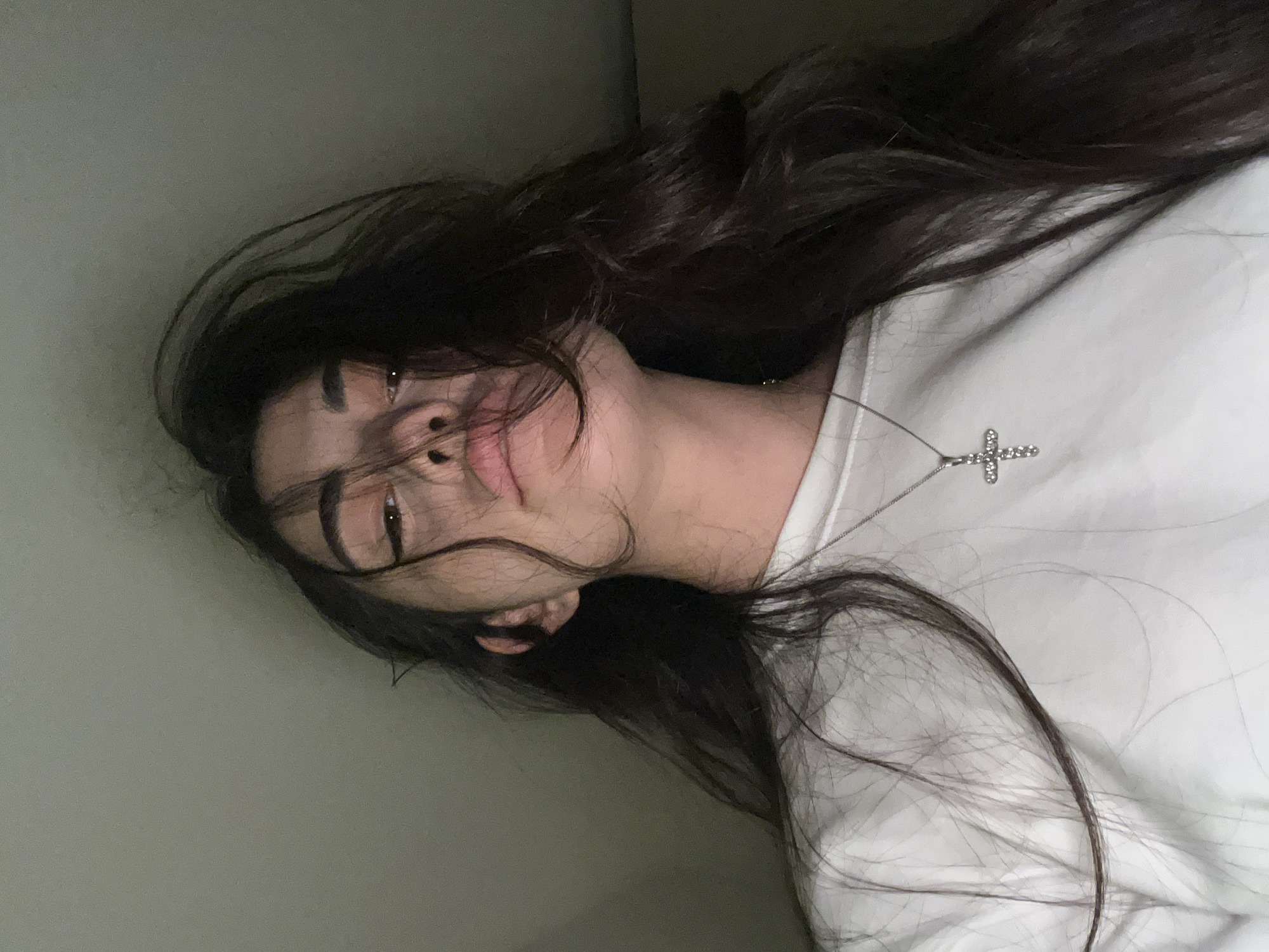 A low-angle, close-up shot captures a young woman from the chest up, her face partially obscured by dark, loose hair. She is wearing a white crewneck sweatshirt and a silver necklace with a small, diamond-encrusted cross pendant. Her eyes are looking upwards, and her lips are slightly parted, creating a pouty expression. The background is a plain, dark gray wall. The scene appears to be indoors, possibly a bedroom or a dimly lit room in Chotěšov, Czechia.