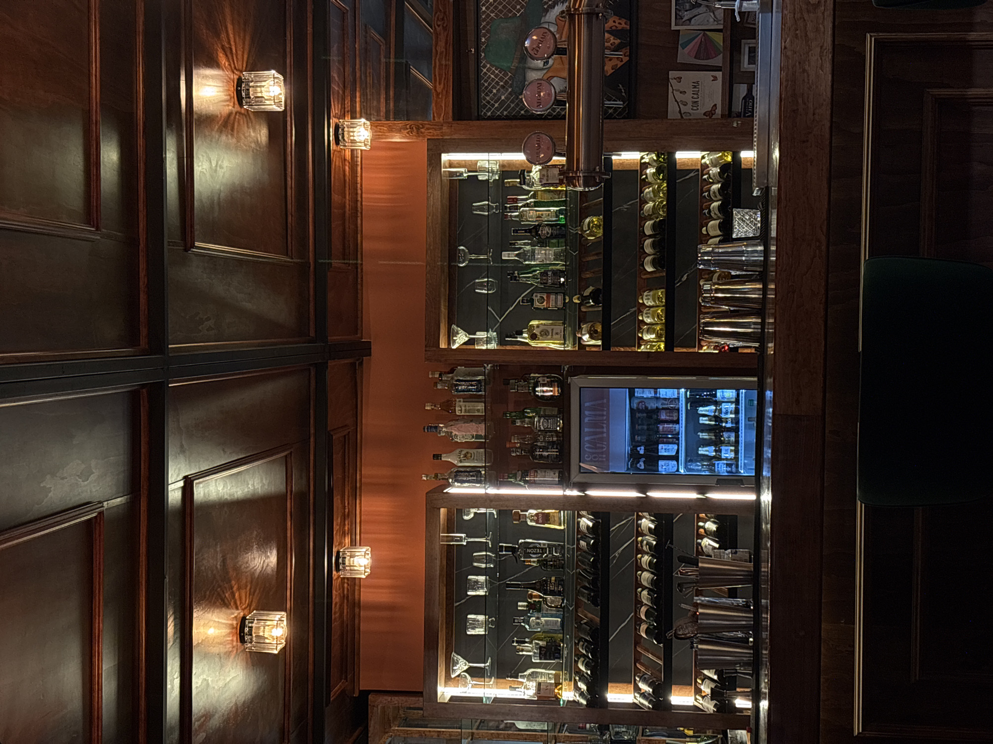 The image captures the interior of a bar in Puebla, Mexico. The scene is dimly lit, with decorative sconces mounted on dark wood-paneled walls that create an ambient glow. A prominent bar structure features illuminated shelves displaying various bottles of alcohol and glassware. A beverage cooler with a selection of beers is visible. The bar area shows a sophisticated and well-stocked environment.