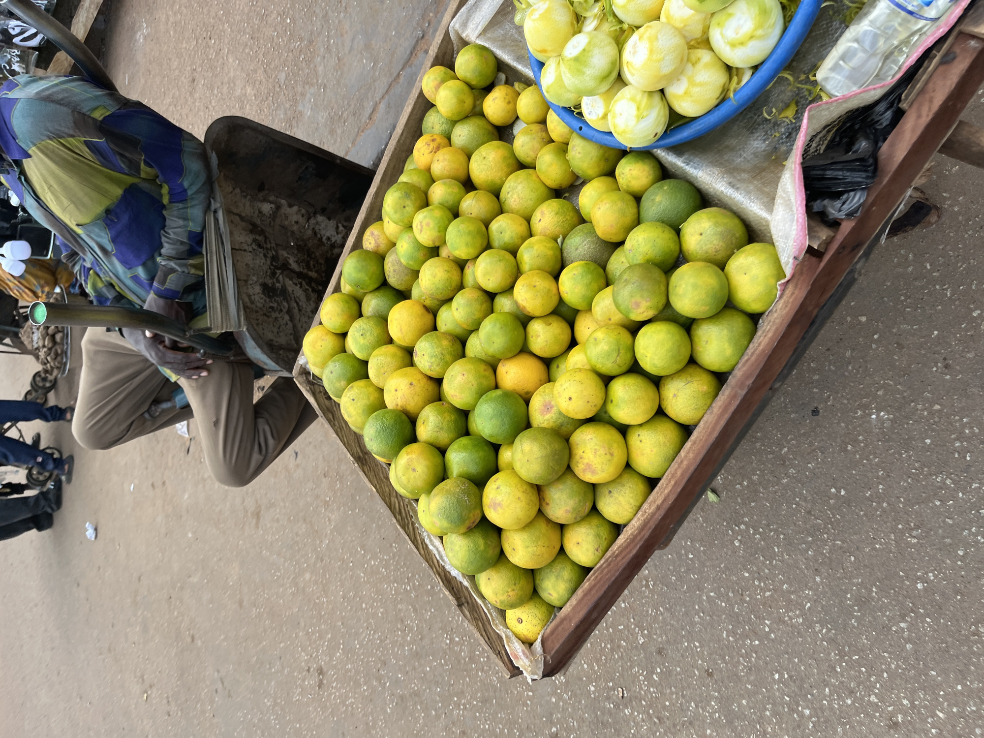 In Bukuru, Nigeria, a man in a checkered shirt and tan pants is seated in a wheelbarrow piled high with oranges. The wheelbarrow's wooden frame is weathered, and it rests on a paved street. To the right of the oranges, a blue plastic bowl contains peeled citrus fruits. In the background, other people are visible, as well as various objects that suggest a market setting. The ground is a dusty, mottled gray, interspersed with small white speckles.