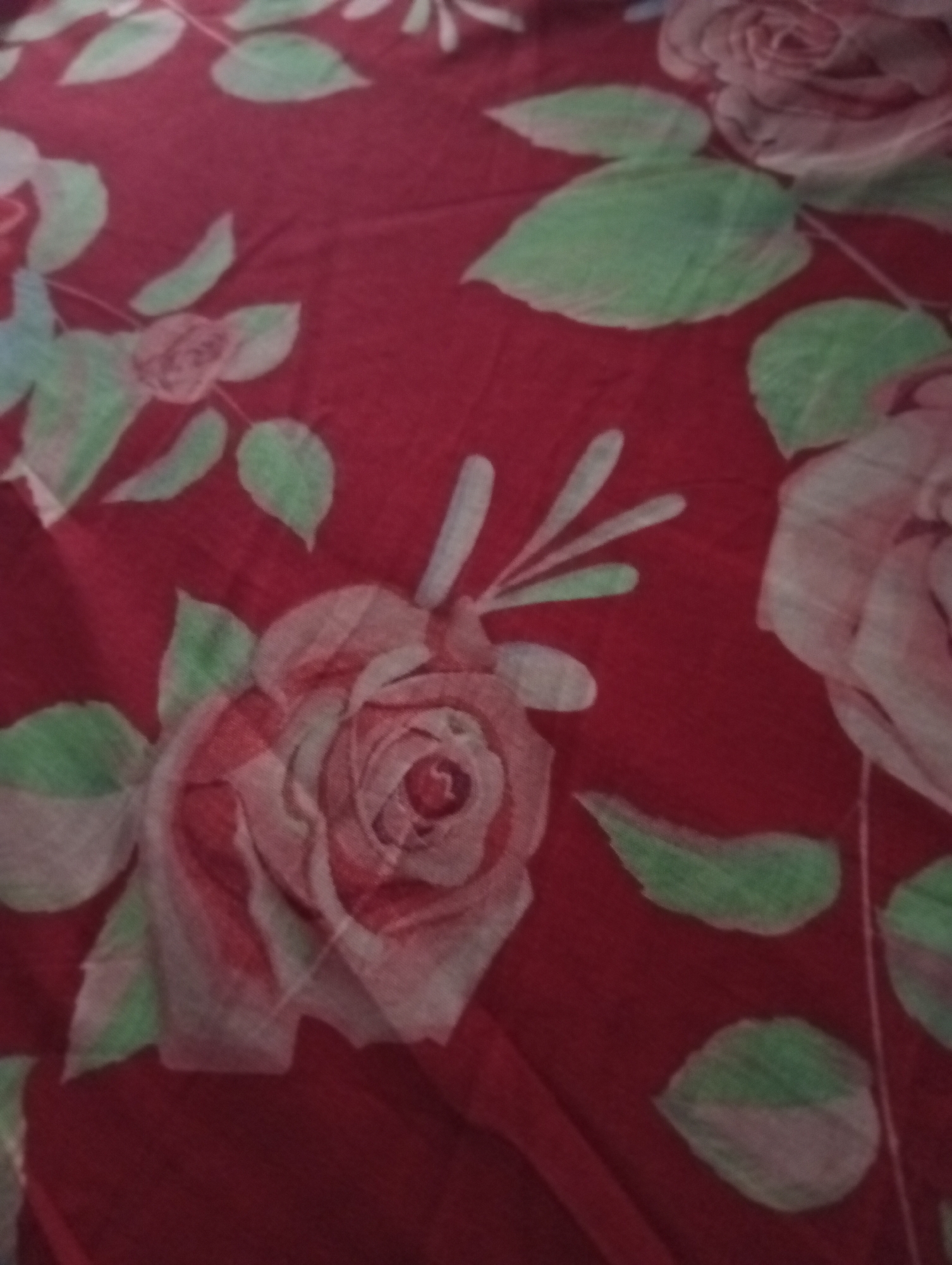 The image displays a close-up of a red fabric with a floral pattern. The pattern consists of roses and leaves in shades of pink, white, and green. The roses are depicted in various stages of bloom, with intricate petal details. The leaves are ovular and have a soft, naturalistic appearance. The fabric appears to be soft and possibly made of cotton or a similar material. The lighting suggests it is an indoor setting, but no specific environmental details or location in Kualu, Indonesia are visible. The image is slightly blurred, making it difficult to discern fine textures or precise details of the pattern. There are no people, objects, or actions depicted in the image.