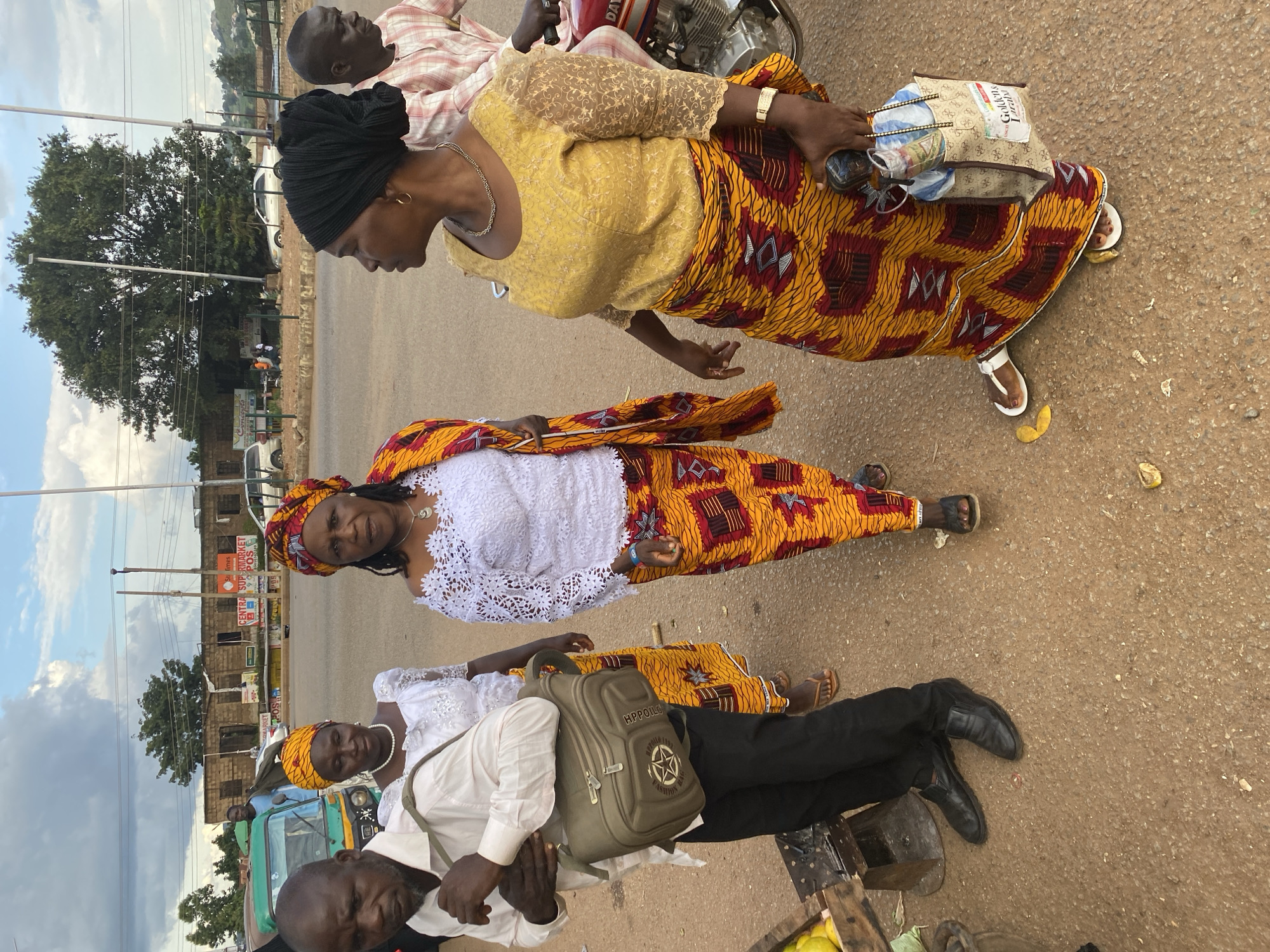 In Bukuru, Nigeria, four individuals, three women and one man, are depicted standing outdoors on a paved road. The man, on the left, wears a light-colored collared shirt, black trousers, and black leather shoes. He carries a khaki messenger bag slung over his right shoulder. The first woman, to his right, is dressed in a white outfit with a pearl necklace. The second woman stands in the center, wearing a white lace top and a vibrant yellow and red patterned wrap skirt. A third woman on the right is adorned in a yellow lace top and a matching yellow and red patterned wrap skirt, with a black head covering. She holds a light-colored patterned tote bag containing various items. A motorcycle is partially visible to her right. The background features buildings, trees, and a cloudy sky. The ground appears to be asphalt, with some scattered debris.