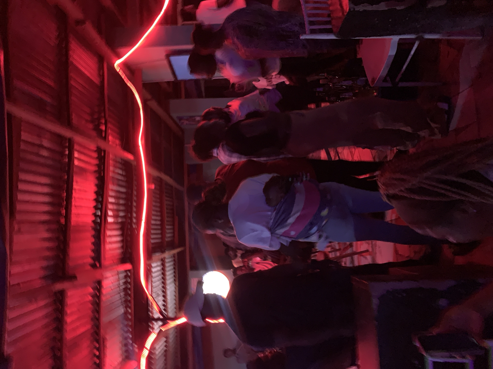 The image depicts a nighttime scene at a location in Kabong, Nigeria. A group of individuals is present, some seated at tables. Red lighting is prominent, illuminating a wall constructed with corrugated metal panels. A glowing bulb is visible, possibly a light source. Interactions between the people are not explicitly discernible from the image, but the context suggests a social gathering. The environment appears to be an indoor or covered space.