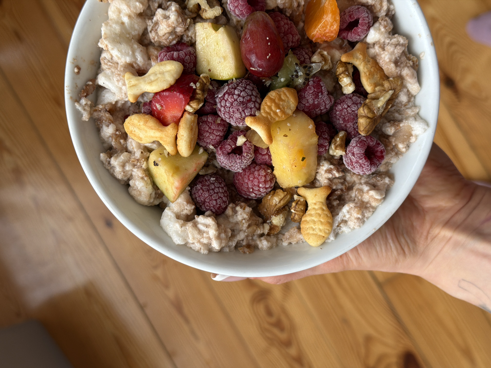 A top-down, close-up shot shows a white bowl filled with a mixture of muesli, raspberries, chopped fruit (apple, plum, tangerine, kiwi), nuts, and fish-shaped crackers. The bowl is held by a hand with a neatly manicured fingernail. The background is a wooden surface, likely a table. The location is not specified, and there is no indication of Budva, Montenegro.