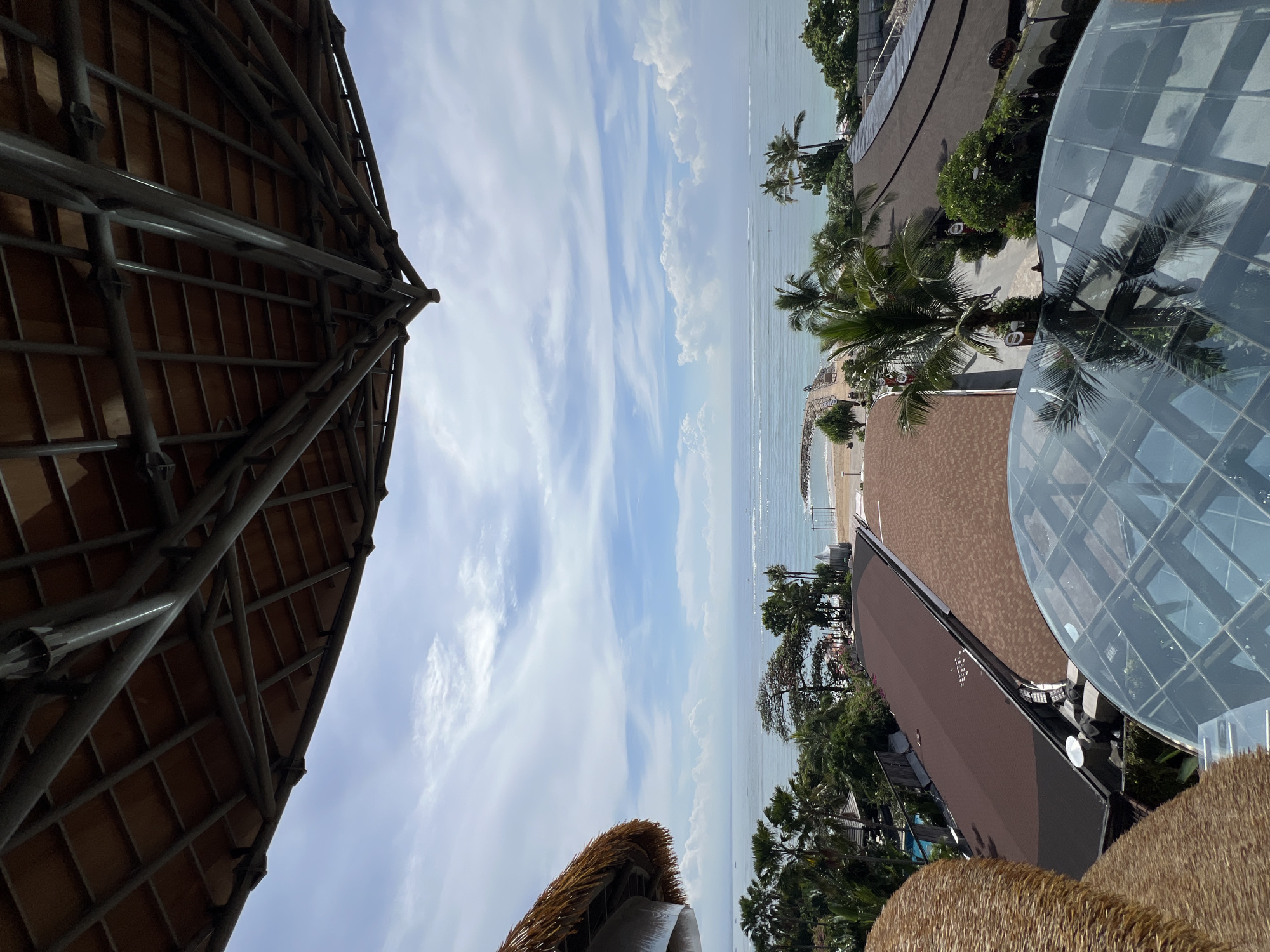 This media file captures an elevated, panoramic view of a coastal resort area, likely in Denpasar, Indonesia. The scene is bathed in bright daylight under a partially cloudy blue sky, featuring long, wispy white clouds.
The upper portion of the image is dominated by the sky and the calm, light-blue ocean stretching to the horizon. A sandy beach is visible along the coastline, dotted with some small structures and leading to lush green foliage and numerous palm trees.
In the lower left foreground, a prominent architectural feature with a large, textured thatched roof supported by dark metal scaffolding or beams takes up a significant portion. This structure appears to be part of a resort building or an elaborate awning.
Towards the lower right, several resort buildings are visible. One particularly notable structure features a large, transparent, hemispherical glass dome, reflecting the surrounding palm trees and the sky. Other buildings nearby have dark brown roofs, and paved paths wind through more tropical trees and plants. The overall impression is one of a tranquil and picturesque tropical vacation destination. No discernible human activity or readable text is visible in the frame.