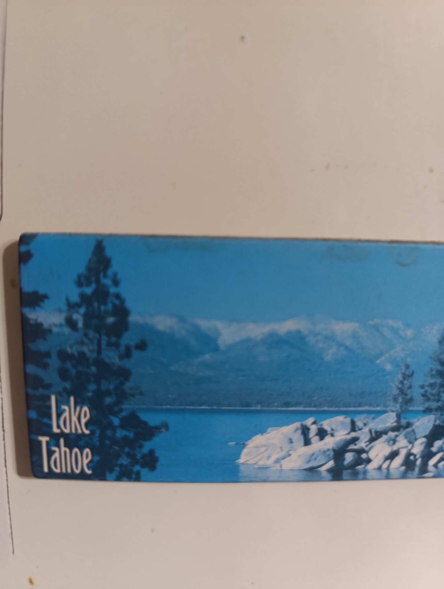 A landscape image depicts a scenic view of Lake Tahoe, with mountains in the background. The foreground features a dark silhouette of pine trees and rocks partially submerged in the water. White text on the bottom left corner reads "Lake Tahoe." The sky is a deep blue, and the water is a vibrant cerulean.