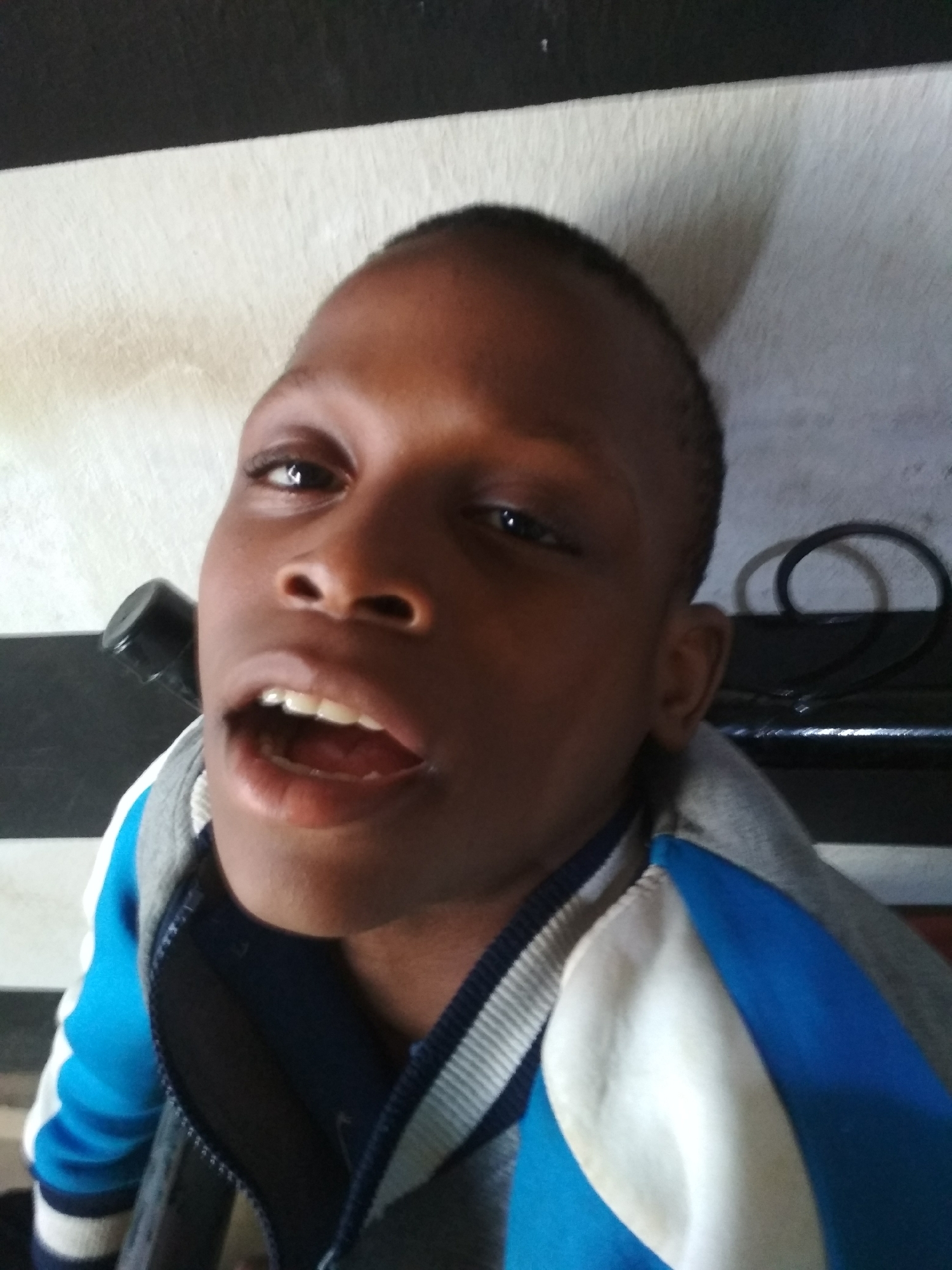 A close-up shot depicts a young Black person with short hair, wearing a blue and gray jacket, with their mouth open. The background features a striped wall of white and dark grey/black. The location is unknown.
