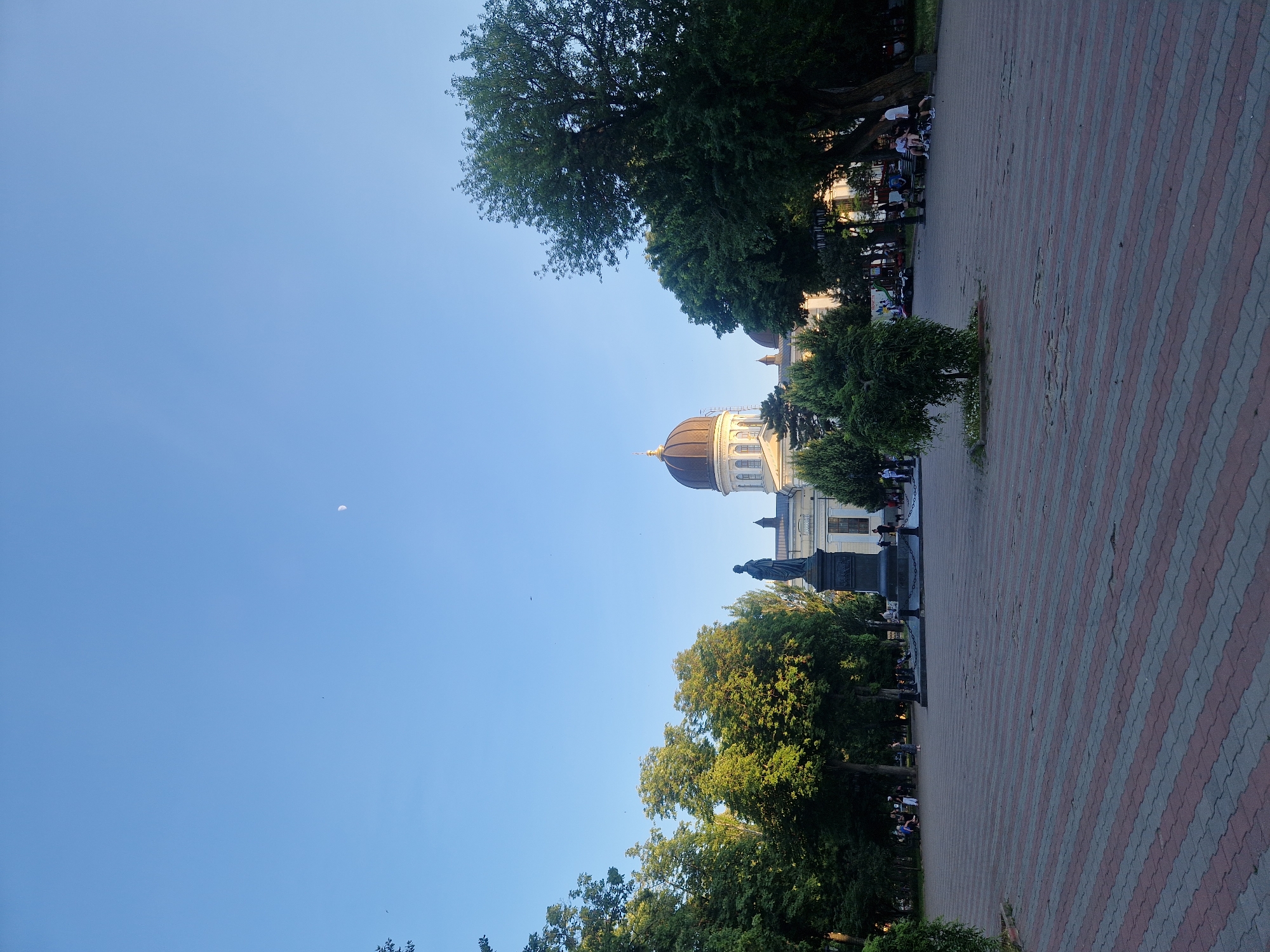 A wide shot shows a park scene with a clear blue sky on the left and a red and gray paved pathway on the right. The pathway is lined with trees and people are visible sitting on benches and walking. In the background, a domed building is visible, suggesting a historical or religious site. A statue of a person in historical attire stands in front of the building, surrounded by trees. The sun appears to be setting, casting a warm glow on the scene, indicated by the long shadows and the golden light on the building. The overall atmosphere is peaceful and relaxed, with people enjoying a pleasant evening in the park. A crescent moon is visible in the sky.