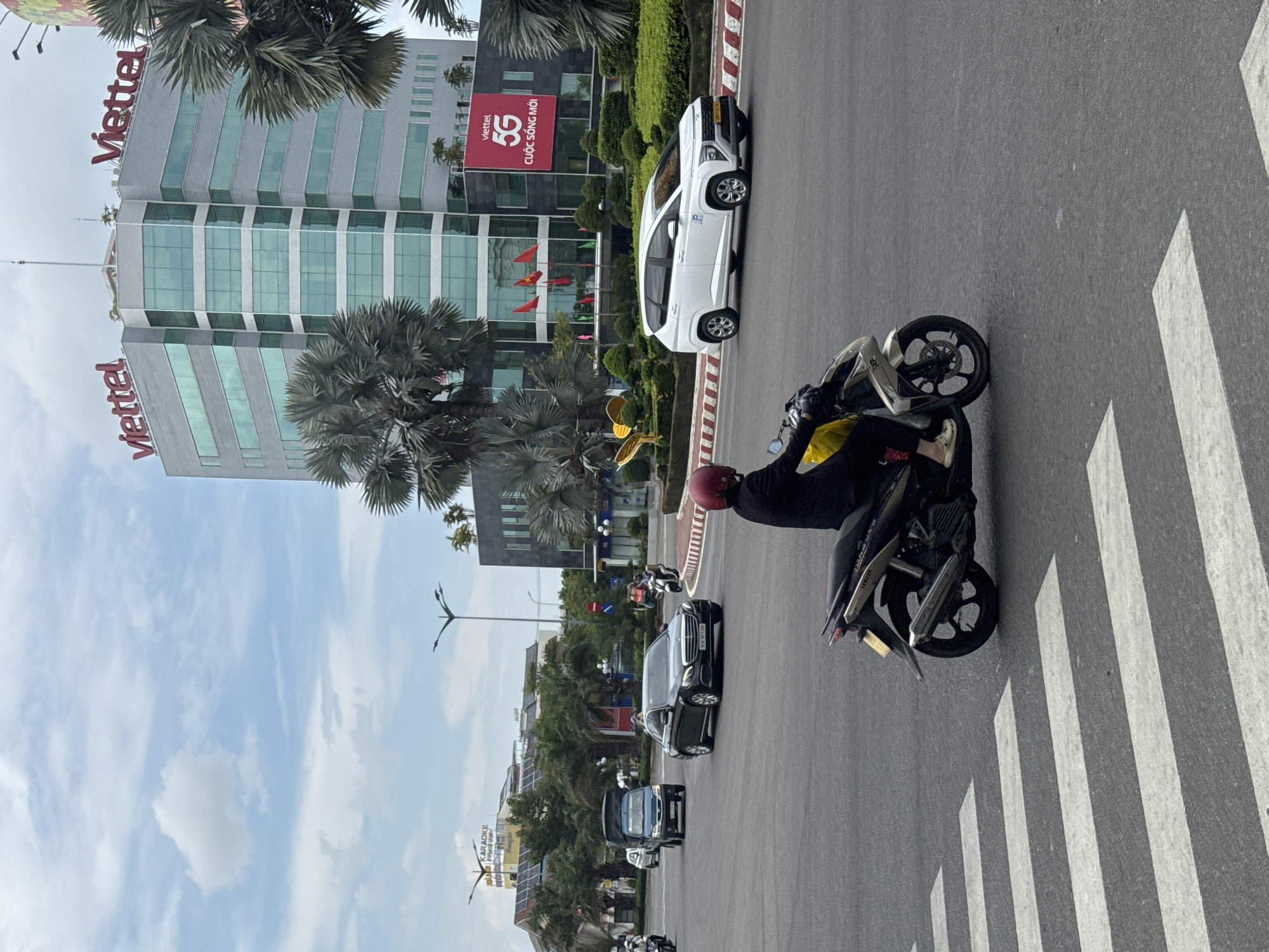 An eye-level, medium shot captures a street scene in Phú Thủy, Vietnam. In the foreground, a person wearing a red helmet and dark clothing rides a motorcycle with a yellow bag attached to the back, crossing a crosswalk. To the right, a white SUV is driving on the road. Behind the SUV, a large, modern building with "Viettel" signage is visible, along with a red banner advertising "Viettel 5G." Palm trees and other greenery are present in the middle ground. Other vehicles, including a dark sedan and a blue truck, are also on the road. The sky is partly cloudy.