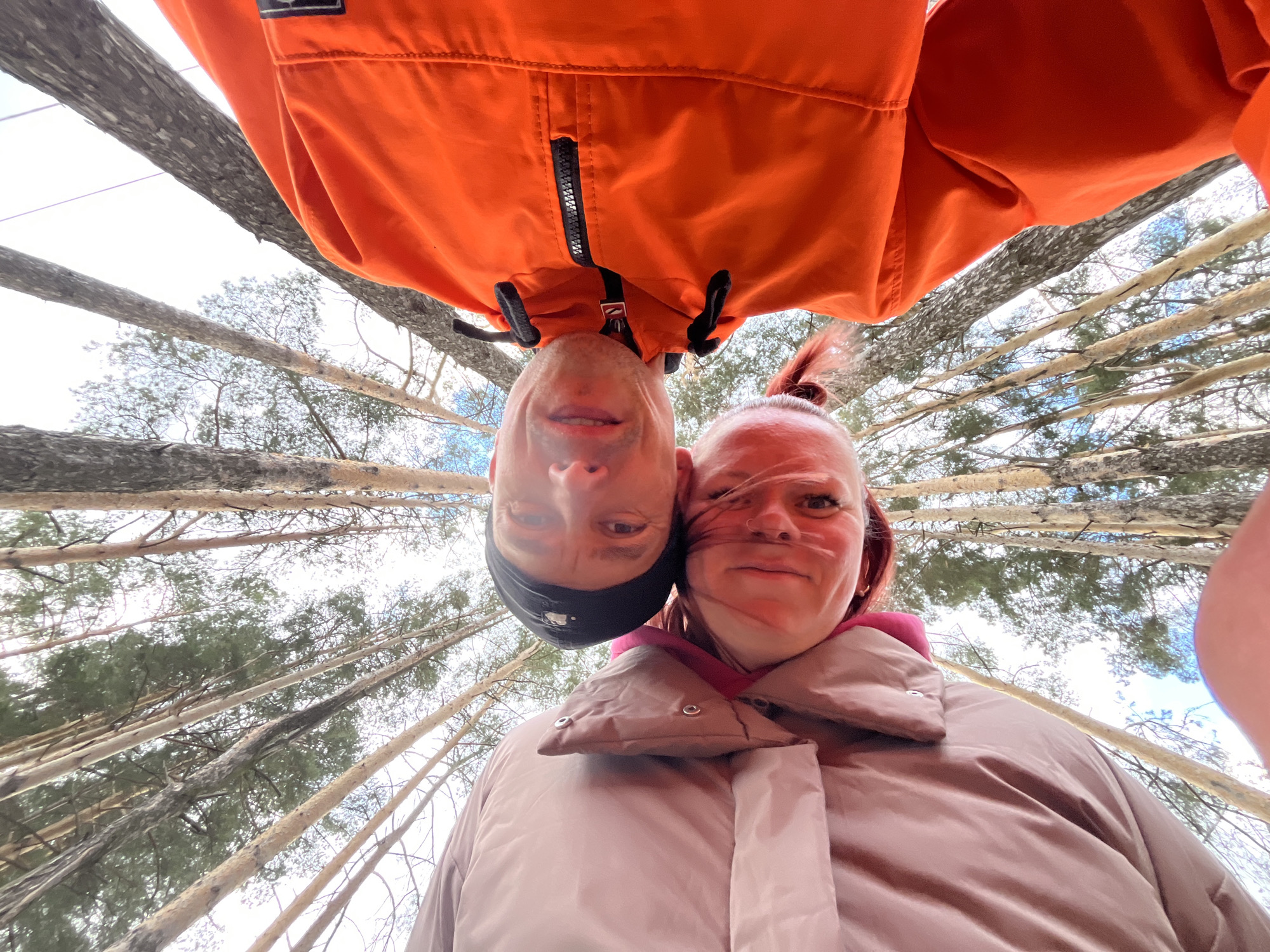 The image captures a low-angle, inverted perspective of two individuals in a forest setting. The camera is positioned looking upwards, with the subjects appearing upside down relative to the viewer.
On the left, a male with a subtle smile wears a vibrant orange jacket featuring a black zipper and a dark cap. On the right, a female with reddish-brown hair tied back, also smiling, wears a light brown or beige padded jacket over a pink layer. Her hair appears gently wind-blown. Both individuals are looking down towards the camera.
The background consists of numerous tall, slender tree trunks, likely pines, reaching towards a bright, possibly overcast sky. Sparse green foliage is visible higher up among the branches. The scene conveys a cheerful outdoor moment, possibly a playful selfie or a photograph taken from a very low vantage point within a forest.
The setting, characterized by the type of trees, is consistent with a natural environment such as those found in Chaikovsky, Russia. The bright conditions suggest the photograph was taken during daytime. No discernible text is visible on clothing or in the background.