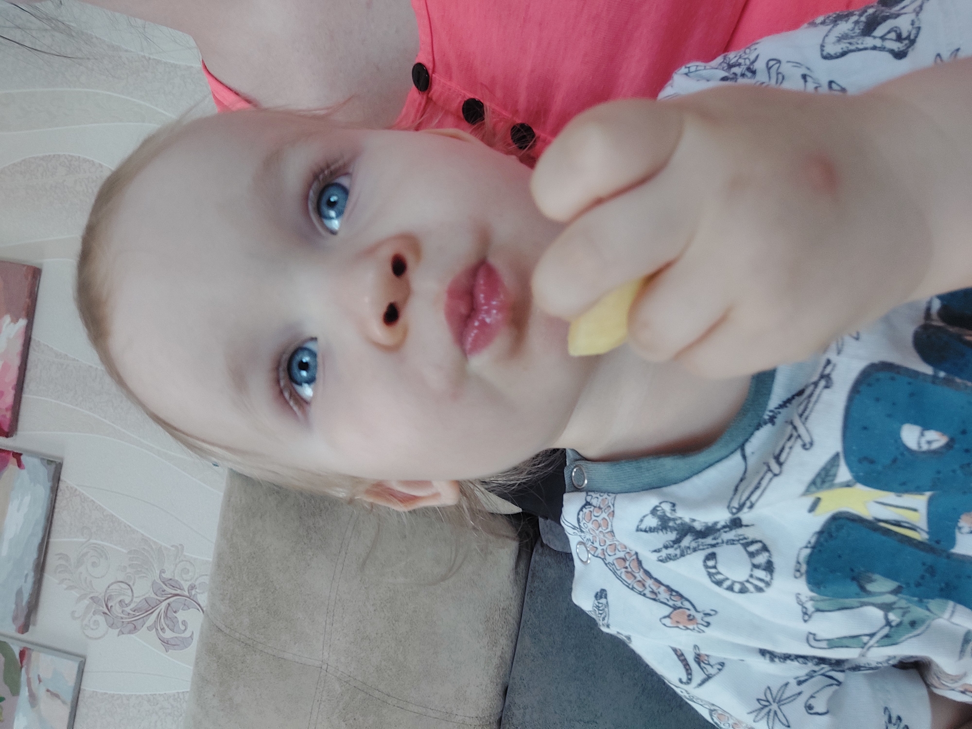 A young child with fair skin, blue eyes, and light blonde hair is eating a yellow food item. The child is wearing a white shirt with blue, yellow, and grey animal patterns and a green trim around the collar. The child's mouth is slightly open, suggesting they are in the act of eating. Part of an adult wearing a coral-colored shirt with black buttons is visible in the background. A patterned wall and part of a neutral-colored sofa are also visible. The location is Большой Букор, Russia.