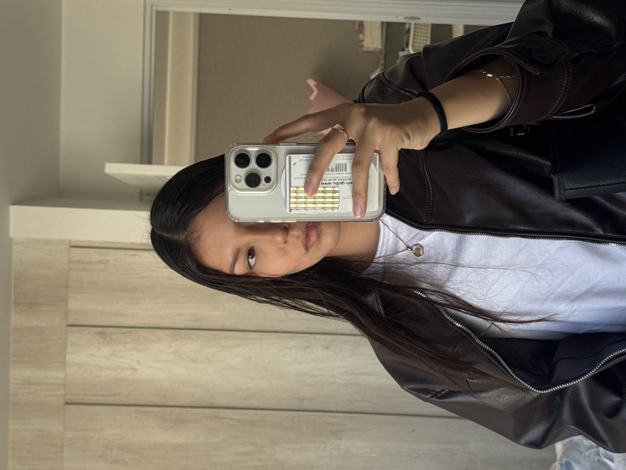 A young woman is captured in a mirror selfie, holding a light-colored smartphone with a clear case that contains several cards, some with golden stripes, visible behind it. The phone's camera module suggests it is a recent iPhone model. She is wearing a dark brown, possibly faux leather, jacket over a white t-shirt, and a delicate silver necklace with a small, light-colored pendant. On her left hand, which holds the phone, she wears a simple ring, and on her right wrist, a thin silver chain bracelet is visible, along with a black elastic hair tie. Her dark hair is long and straight, and she looks directly towards the phone's lens with a neutral expression.
The setting appears to be indoors, likely a room with light-toned walls and possibly wooden paneling or floor visible in the background reflection. A faint pink object is reflected in the mirror behind her. The lighting is bright, suggesting daytime. The entire image is rotated 90 degrees counter-clockwise.
Visible text on the cards within the phone case is inverted and blurry, making it largely indistinguishable. However, one card seems to display a URL starting with "WWW.A..." and another card shows lines of text and what appears to be a barcode, possibly indicating an identification or credit card. This scene takes place in San Francisco Ocotlán (Ocotlán), Mexico.