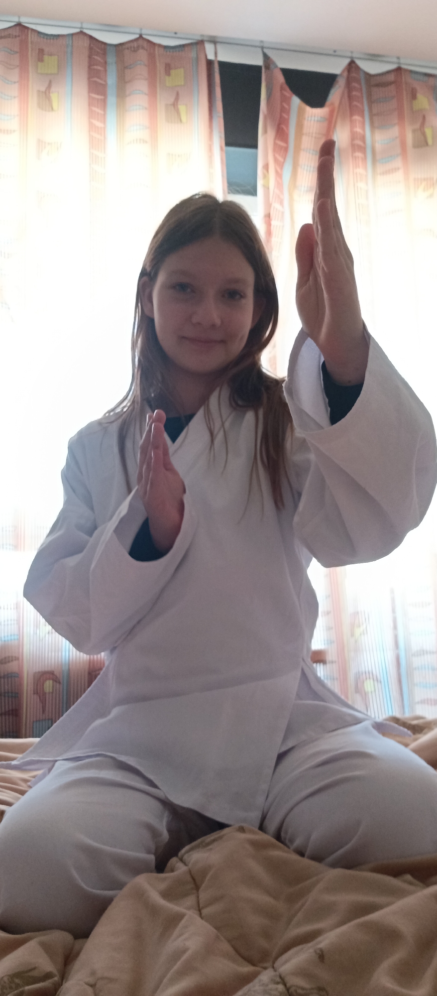 A young person is kneeling on a bed, wearing a white martial arts uniform. The person is facing the viewer and has their hands raised in front of them. The background includes a window with curtains. The location is in a city named Unknown, in an unknown location. The bed is covered with a brown comforter.