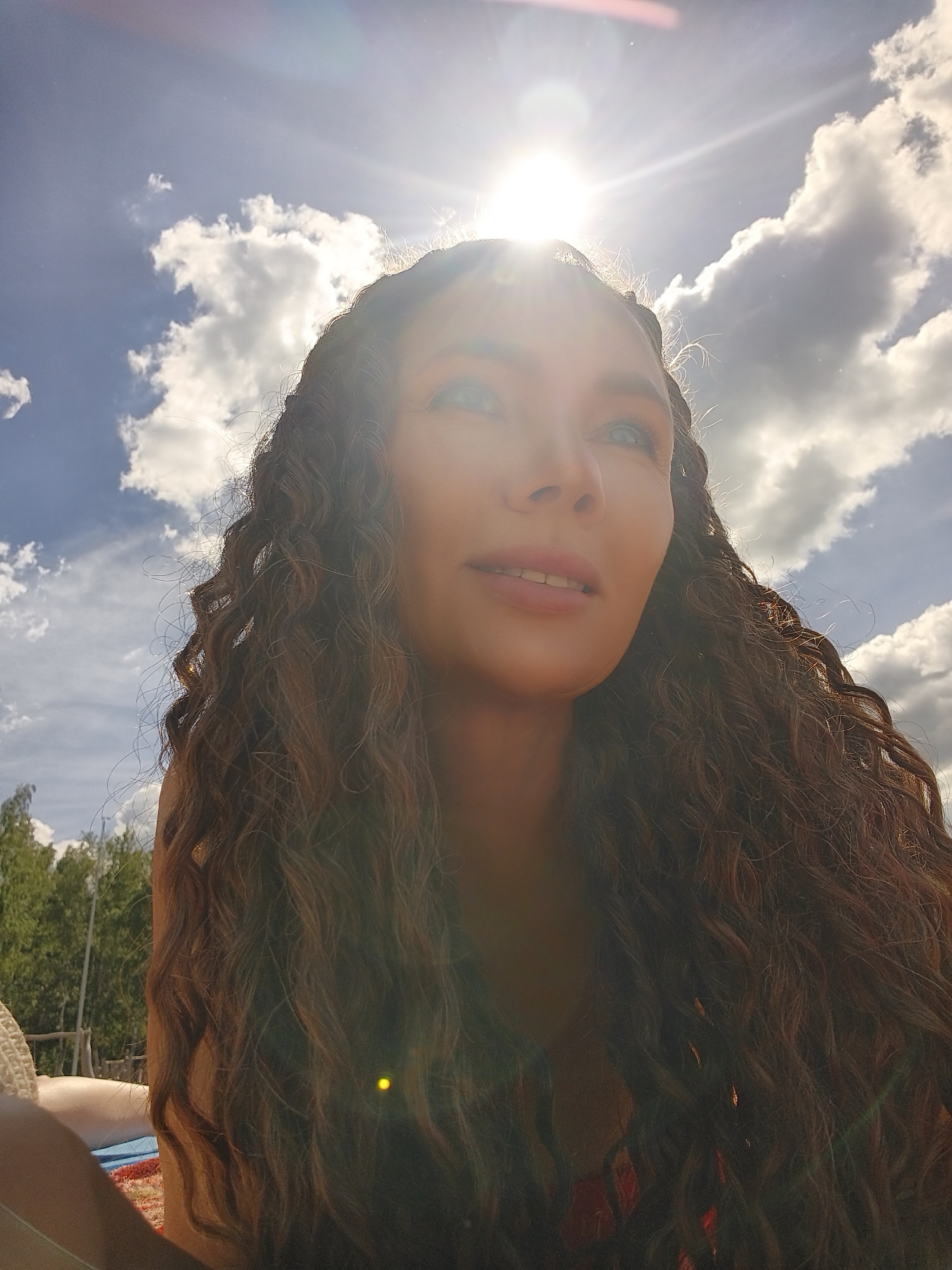 In a low-angle, outdoor, medium shot, a woman with long, curly brown hair is captured against a bright sky with scattered clouds and the sun shining intensely. The sun's rays create a lens flare, obscuring the upper portion of the image and parts of the woman's face. Her head is tilted upwards and to the right, with her eyes gazing in that direction. Her mouth is slightly open, revealing her teeth. Her skin appears tanned. Behind her, a blurry background shows green trees and a wooden structure, suggesting a natural or recreational setting. The woman is wearing a dark garment, possibly a bathing suit top, visible at the bottom of the frame. The lighting is harsh and backlit.