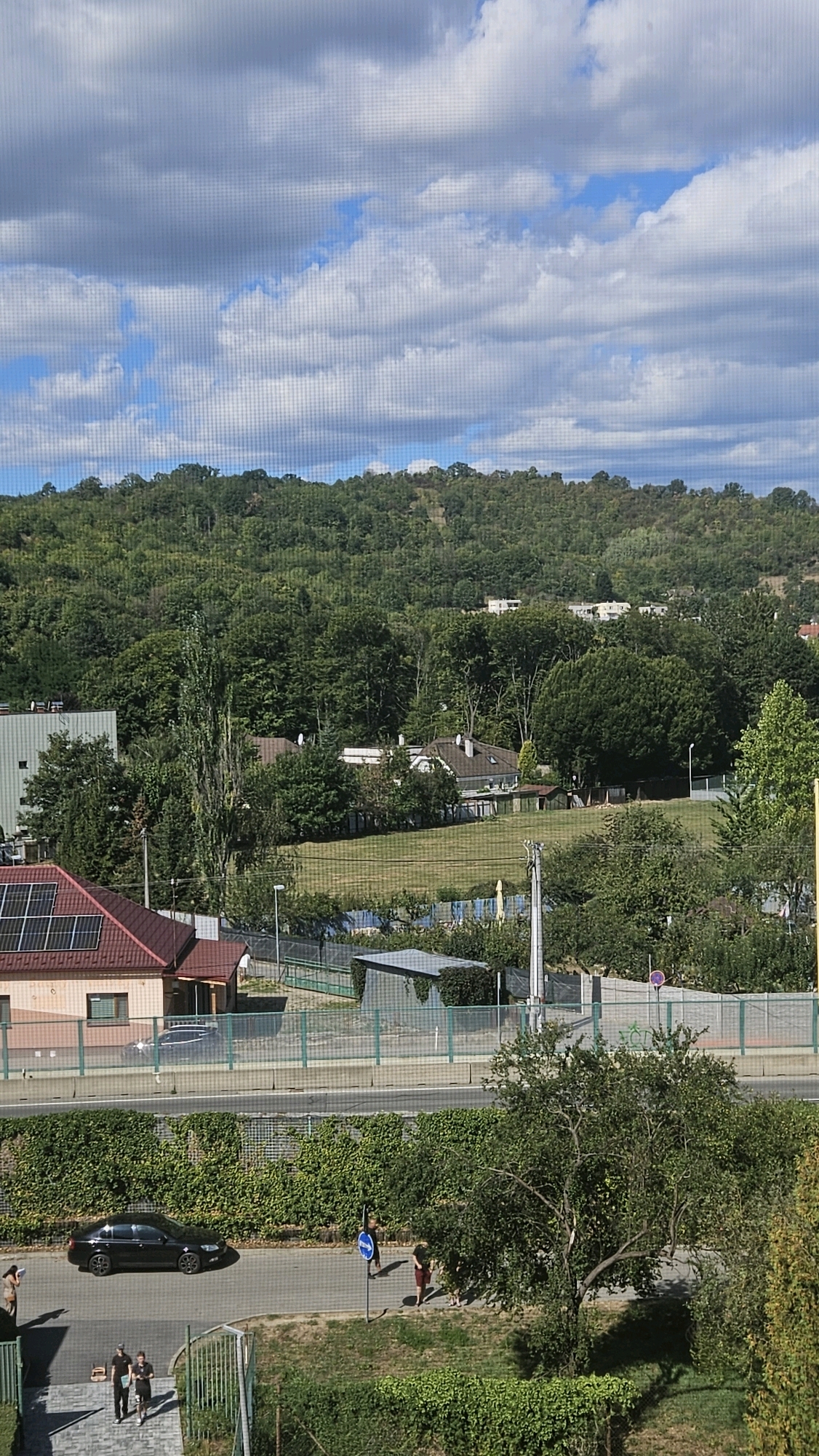 The image shows a suburban or semi-rural residential area with a verdant hillside in the background. In the foreground, there is a paved road with a black car driving on it. Two individuals are walking on the sidewalk next to the road, one wearing a dark dress and the other in dark shorts and a t-shirt. Another individual in a brown dress is standing further down the sidewalk. Across the road, there is a fence and a dense green hedge. Beyond that, there are several buildings, including a house with a red roof and solar panels, and a larger industrial or commercial building. Further back, a neatly mown grassy field is visible, bordered by trees and fences. In the distance, houses are scattered on the densely wooded hillside under a partly cloudy sky. The scene appears to be in Prešov, Slovakia, as indicated by the presence of typical European residential architecture and landscaping. The overall impression is one of a sunny day in a well-maintained community.