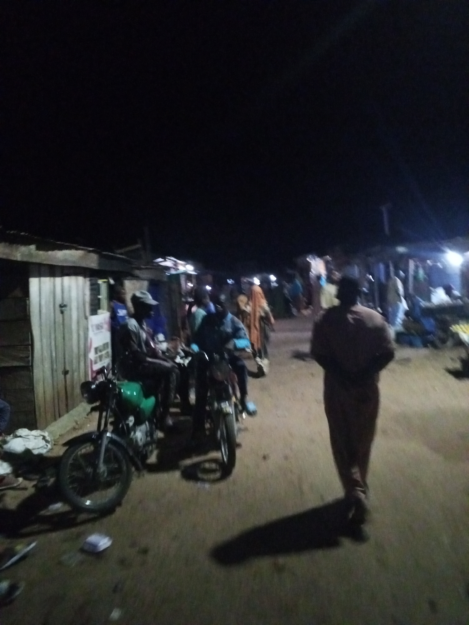 At night in Lokoja, Nigeria, an unpaved street scene shows a dimly lit area populated with people. Two motorcycles are parked on the left side of the frame; one is green. Several individuals are either seated on the motorcycles or standing nearby. A man in a brown outfit walks away from the viewer down the street. Stalls and other pedestrian traffic are visible in the background. The light appears to be artificial, coming from the shops or stands. A building is on the left with a sign on it. The ground has debris.