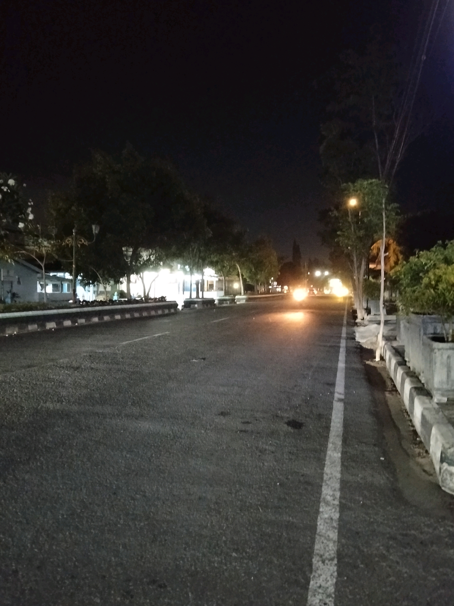 The image depicts a street scene at night in Kliwonan, Indonesia. An asphalt road with a white lane marker runs through the center of the frame, angling towards the background. On the left side of the road, a low concrete barrier with alternating black and white rectangular blocks delineates the edge. Beyond the barrier are trees, some with white flowers, and illuminated buildings. On the right side of the road, a similar concrete barrier borders the street, with planters containing green foliage. Several streetlights are visible, casting a warm glow. In the distance, the headlights of a vehicle create a bright, blurry streak of light on the road. The sky is dark, indicating it is nighttime.