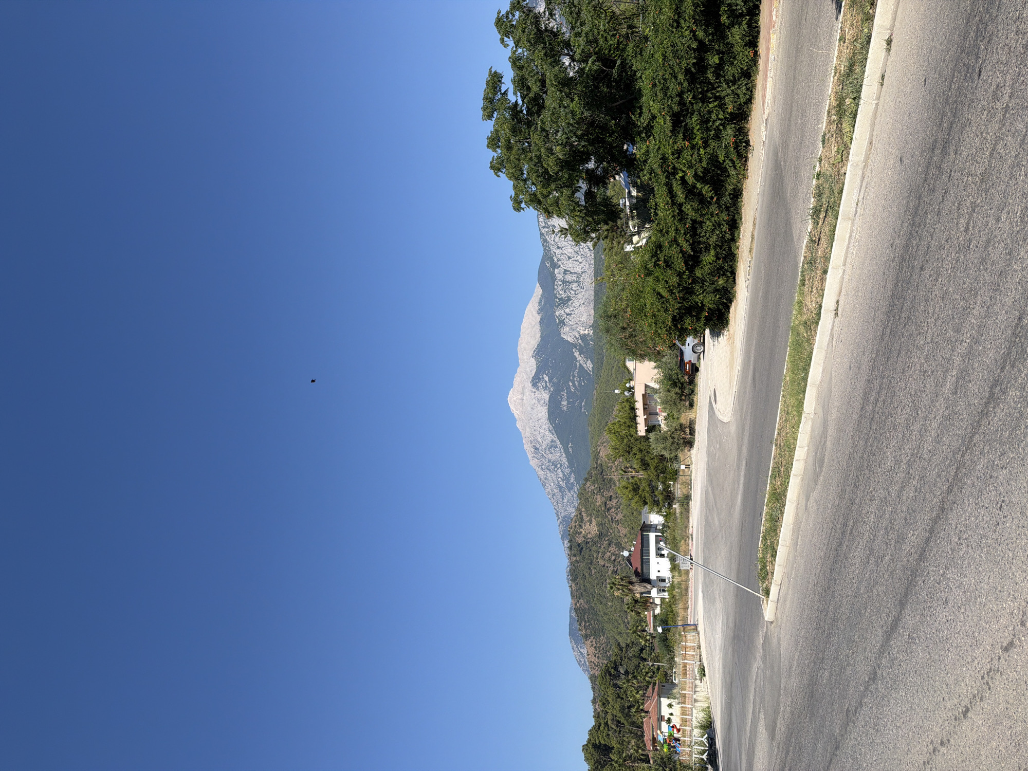 The image captures a scenic view of a road leading towards a mountainous landscape under a clear blue sky. The foreground features an asphalt road with a concrete curb and a patch of dry grass. To the left, a white fence separates the road from a small playground with colorful structures. Behind the fence, several houses with red-tiled roofs are visible, interspersed with palm trees and other greenery.
Further in the background, a dense forest covers the lower slopes of the mountains, which rise majestically into the distance. The peaks of these mountains are bare and rocky, appearing light in color, possibly due to snow or geological composition. A single bird is visible as a small black silhouette against the vast expanse of the sky, near the center top of the image. On the right side of the image, a large, lush green tree dominates the midground, partially obscuring some buildings and the mountain view. A silver car is parked on the side of the road, partially visible behind this tree. The overall impression is one of a tranquil, semi-rural setting in or near Kemer, Turkey, with a striking natural backdrop.