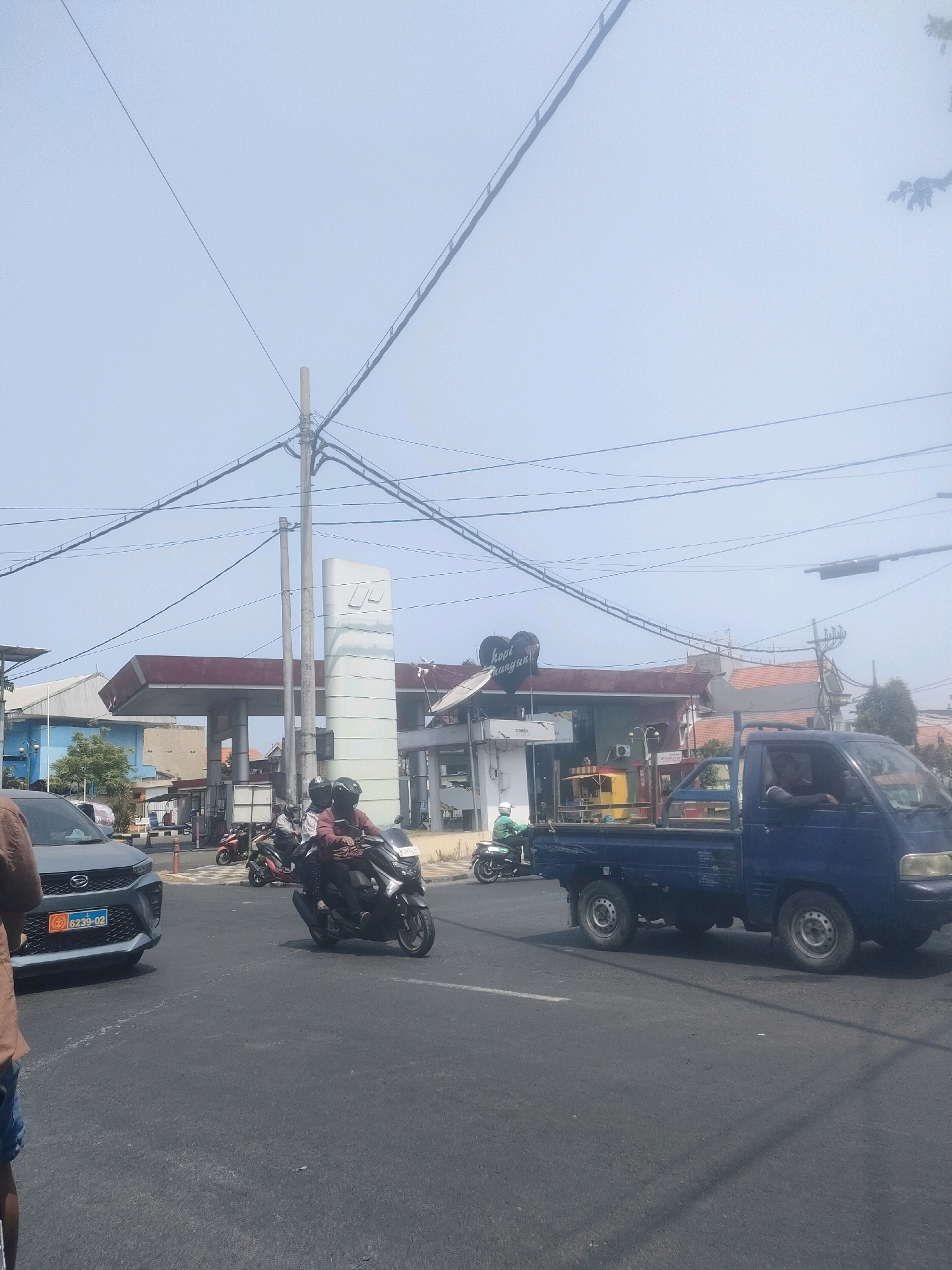 The image depicts a street scene in Surabaya, Indonesia, under a clear, hazy sky. In the foreground, a dark gray sedan with a blue license plate reading "6239-02" is partially visible on the left. To the right of the sedan, two individuals on a black scooter wear helmets, with the driver sporting a red shirt and the passenger a dark top. Further into the mid-ground, a blue pickup truck drives past. Behind the vehicles, a gas station with red and white branding and a prominent white pillar is visible. Adjacent to the gas station, a building with signage reading "kopi unyuguan" in a heart shape is present. Several power lines crisscross the sky above the scene. The road appears to be an intersection with visible white lane markings. A person wearing a brown shirt and blue shorts is partially visible in the extreme left foreground.