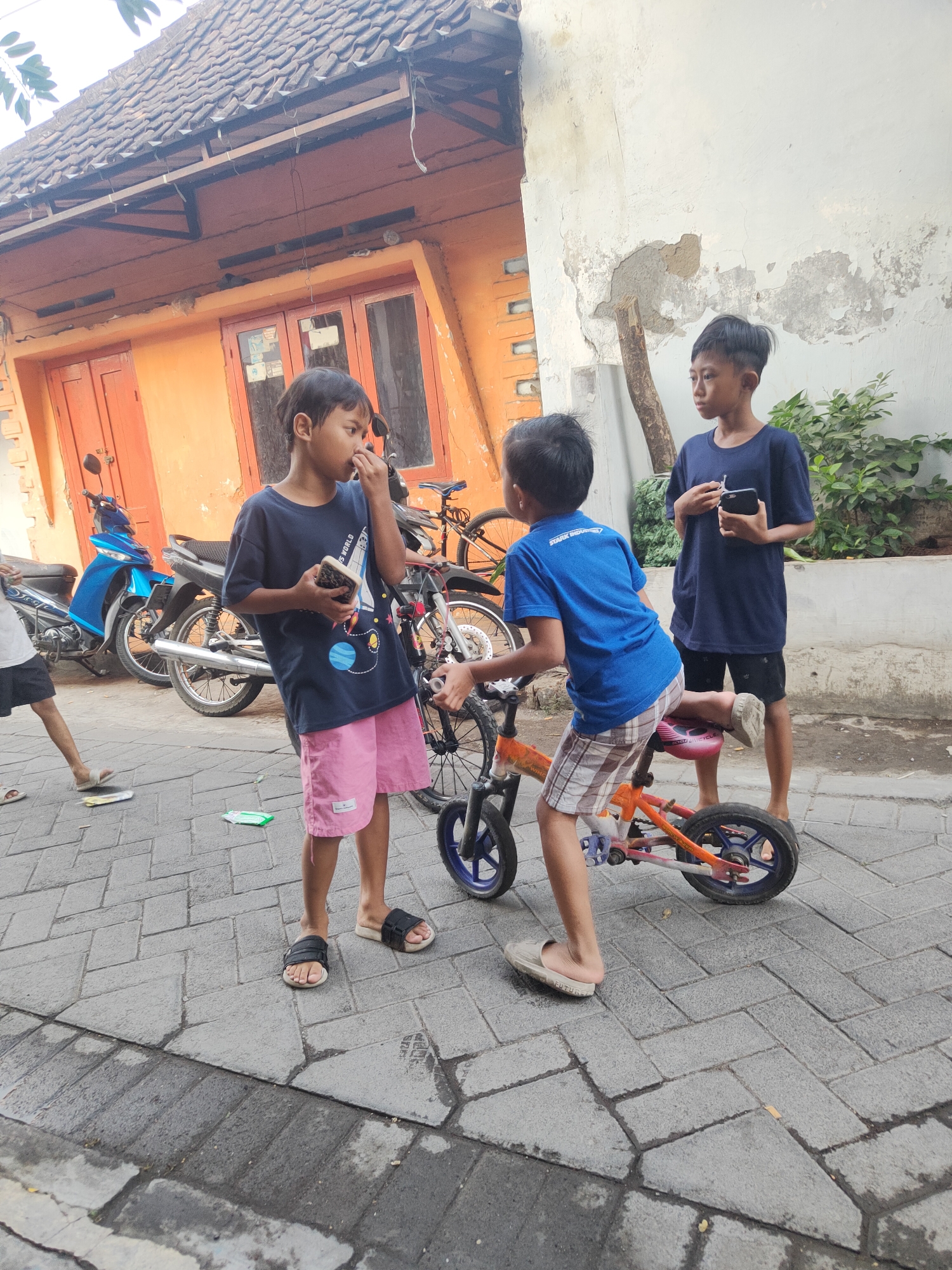 In Kauman, Indonesia, three young boys are interacting on a paved street in front of an orange building. One boy, wearing a dark blue t-shirt and pink shorts, stands with his phone in his hand, bringing his fingers to his nose. To his right, another boy in a blue t-shirt and checkered shorts sits on a small, orange bicycle. He is looking forward and has one foot on the pedal, appearing to be about to push off or dismount. A third boy, in a dark blue t-shirt and black shorts, stands behind the boy on the bicycle, holding a black smartphone. Several motorcycles are parked behind the first boy, and a portion of the orange building with red doors is visible. The environment appears to be a residential area with a textured, brick-like pavement and some greenery in the background.