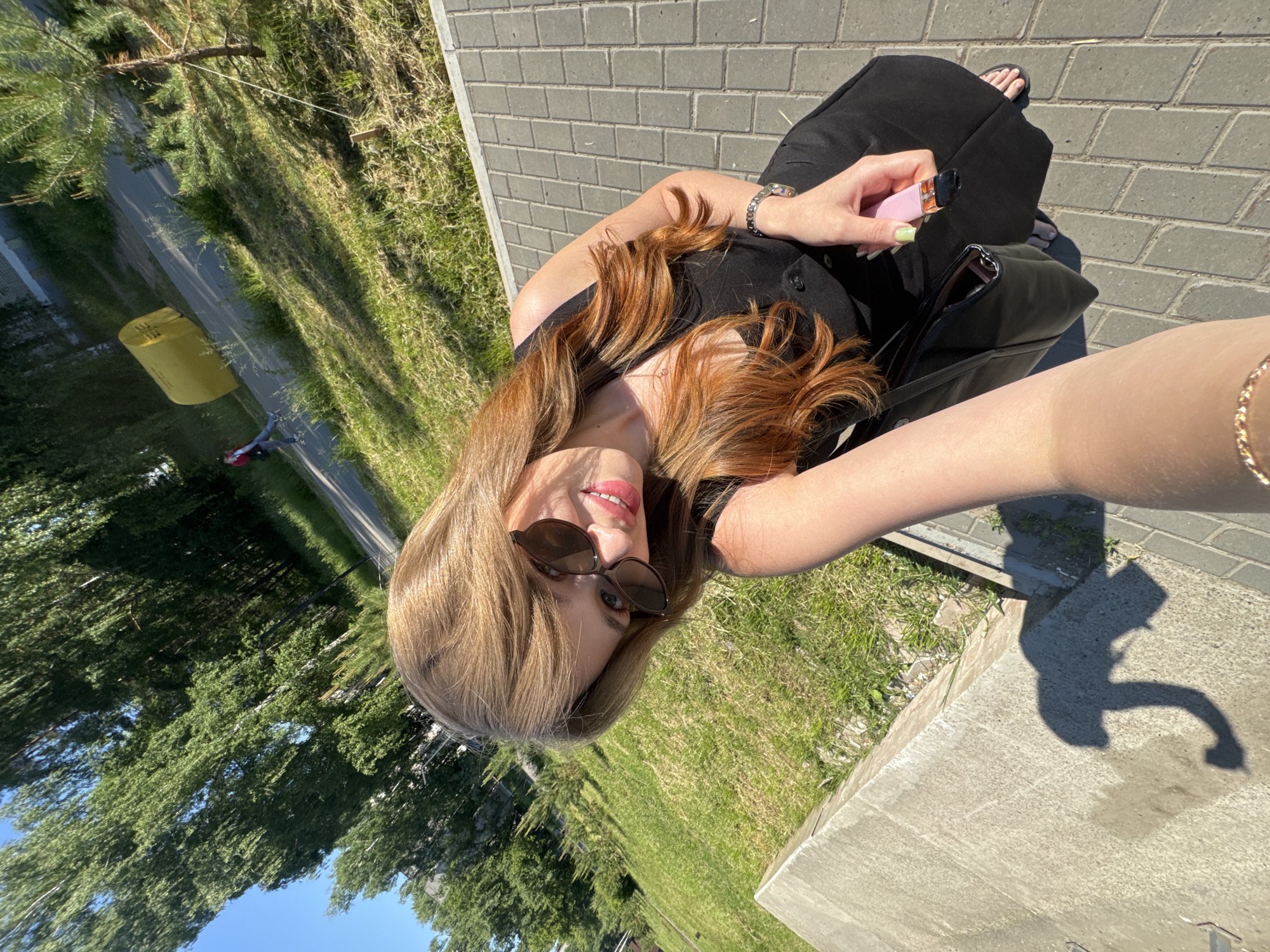 The image shows a woman taking a selfie outdoors in Chaikovsky, Russia. She is wearing sunglasses, a black dress, and holding a pink vape pen in her right hand. Her left arm is extended, holding the camera. She has long, wavy brown hair with reddish undertones. The background consists of a paved walkway, green grass, trees, and a blue sky. A yellow cylindrical object is visible in the distance on the right side of the image, and a person is walking on a path in the background. The lighting suggests it is a sunny day.