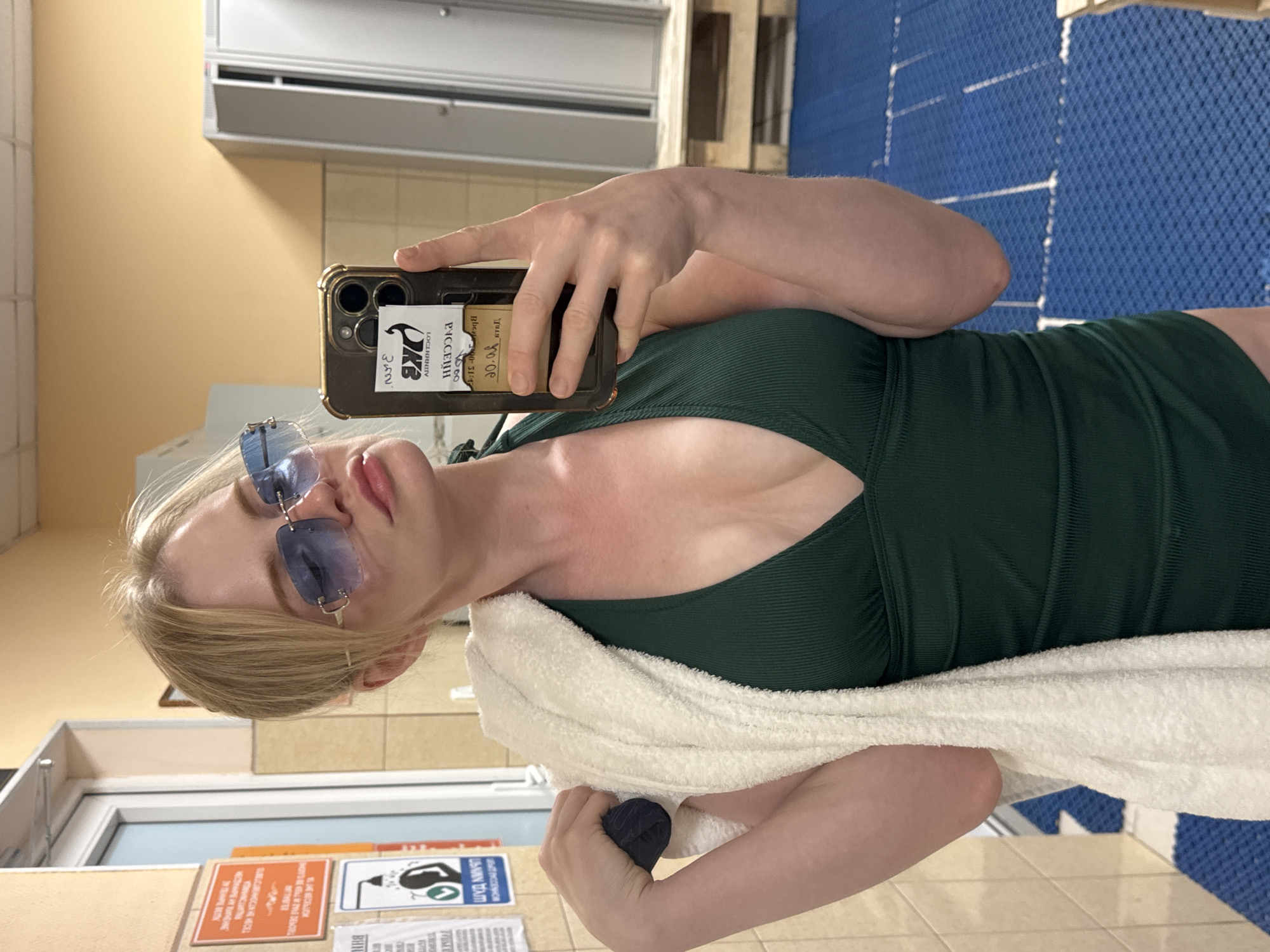 The image shows a young blonde woman with blue-tinted sunglasses and a green swimsuit taking a selfie in what appears to be a locker room. She is holding a white towel over her left shoulder. The locker room has tiled walls, blue matting on the floor, and metal lockers in the background. There are several signs on the wall to her left, one of which appears to be a warning sign with pictograms. A phone with a black case and a sticker is visible in her right hand, as she uses it to take the photo. The overall setting suggests a public facility like a swimming pool or gym locker room. The yellow accent wall and tiled walls are consistent with common locker room aesthetics in many public facilities.