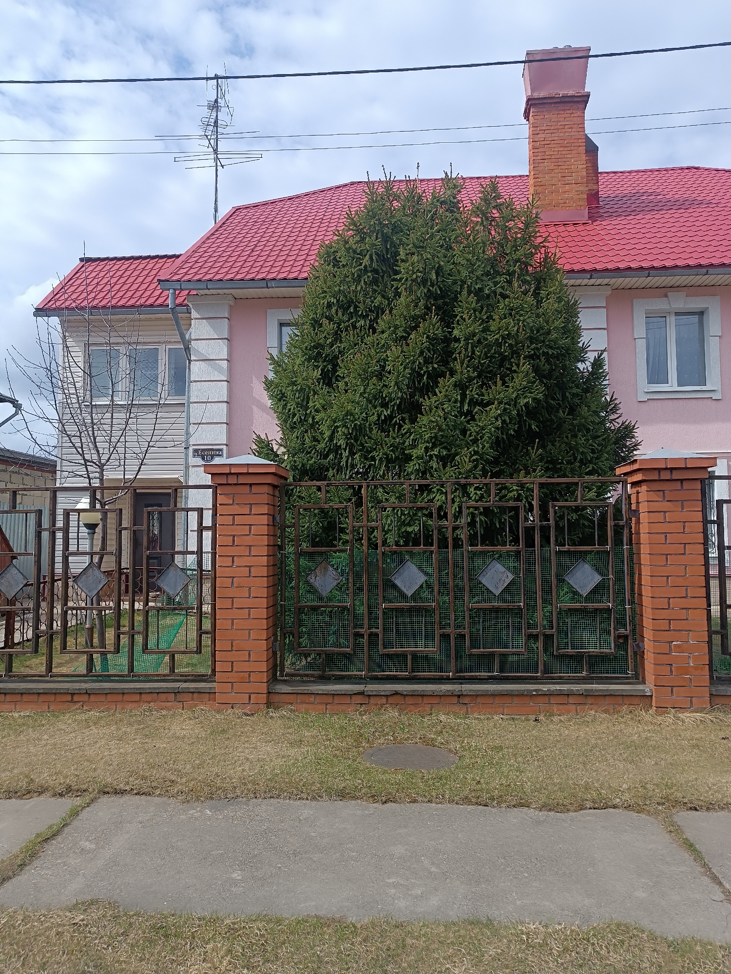 The image shows a two-story house in Chaikovsky, Russia. The house has a pink facade, a red roof, and a prominent brick chimney. A large, dark green coniferous tree obscures part of the front of the house and the iron fence with brick pillars runs along the front property line. The grass in the yard is brown and patchy. A concrete sidewalk is in the foreground. An antenna is visible on the roof. A street sign "Есенина 10" is visible on the corner of the house.