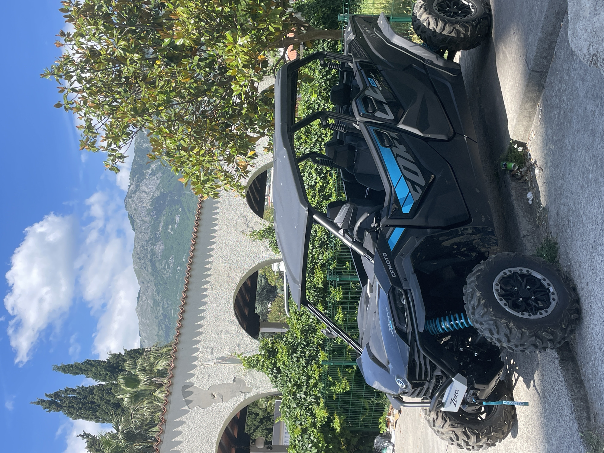 A CFMOTO ZFORCE all-terrain vehicle sits parked on a sidewalk next to a building with white walls and arched doorways in Budva, Montenegro. Vegetation is visible around the building and adjacent to the vehicle. In the background, mountains are visible under a blue sky with scattered clouds.