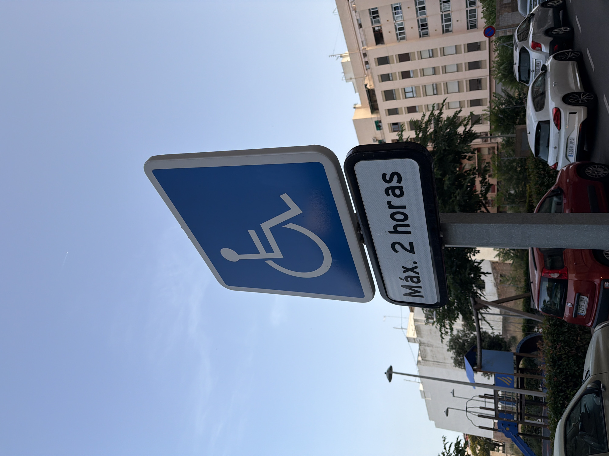 The image shows a street scene in Castelló De La Plana, Spain, featuring a disabled parking sign. The sign is blue with a white wheelchair symbol and a white border. Below it is a rectangular sign with black text on a white background that reads "Máx. 2 horas" (Maximum 2 hours). Several cars are parked on the street, including a red Fiat, a white Volkswagen Scirocco, and a silver SUV. In the background, there is a playground with a slide and swings, and a multi-story apartment building. The sky is clear and blue with a few wisps of clouds. The scene appears to be captured during daylight hours.
