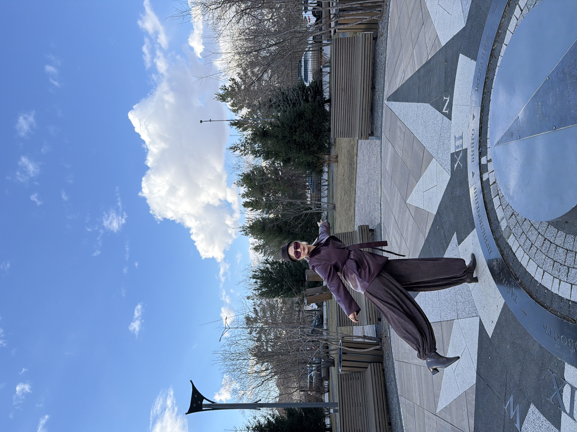 A person, appearing to be a woman, is captured in a dynamic pose on a bright, sunny day in what appears to be a public square or park in Krasnoyarsk, Russia. She is dressed in a dark purple or brown belted jacket, wide-legged patterned trousers, and dark boots with a distinctive heel. She also wears sunglasses and a matching hat. Her left arm is outstretched, pointing to the side, while her right leg is lifted, suggesting an active or posed stance.
She is standing on a large, decorative ground compass rose or sundial, constructed from contrasting light and dark grey stone tiles. Visible Cyrillic text on the metal ring of the compass includes "ЖЕЛАНИЕ" (Desire), "БИНГО" (Bingo), and the city name "КРАСНОЯРСК" (Krasnoyarsk). The Roman numeral "XII" and a large letter "Z" are also discernible on the tile work.
The background features a mix of bare-branched deciduous trees and dark green evergreen trees, indicating perhaps early spring or late autumn. Several wooden park benches are visible along a paved path. Further in the distance, cars are parked along a street, with more trees and urban structures beyond. The sky is bright blue with scattered white cumulus clouds, suggesting clear and pleasant weather during the daytime.