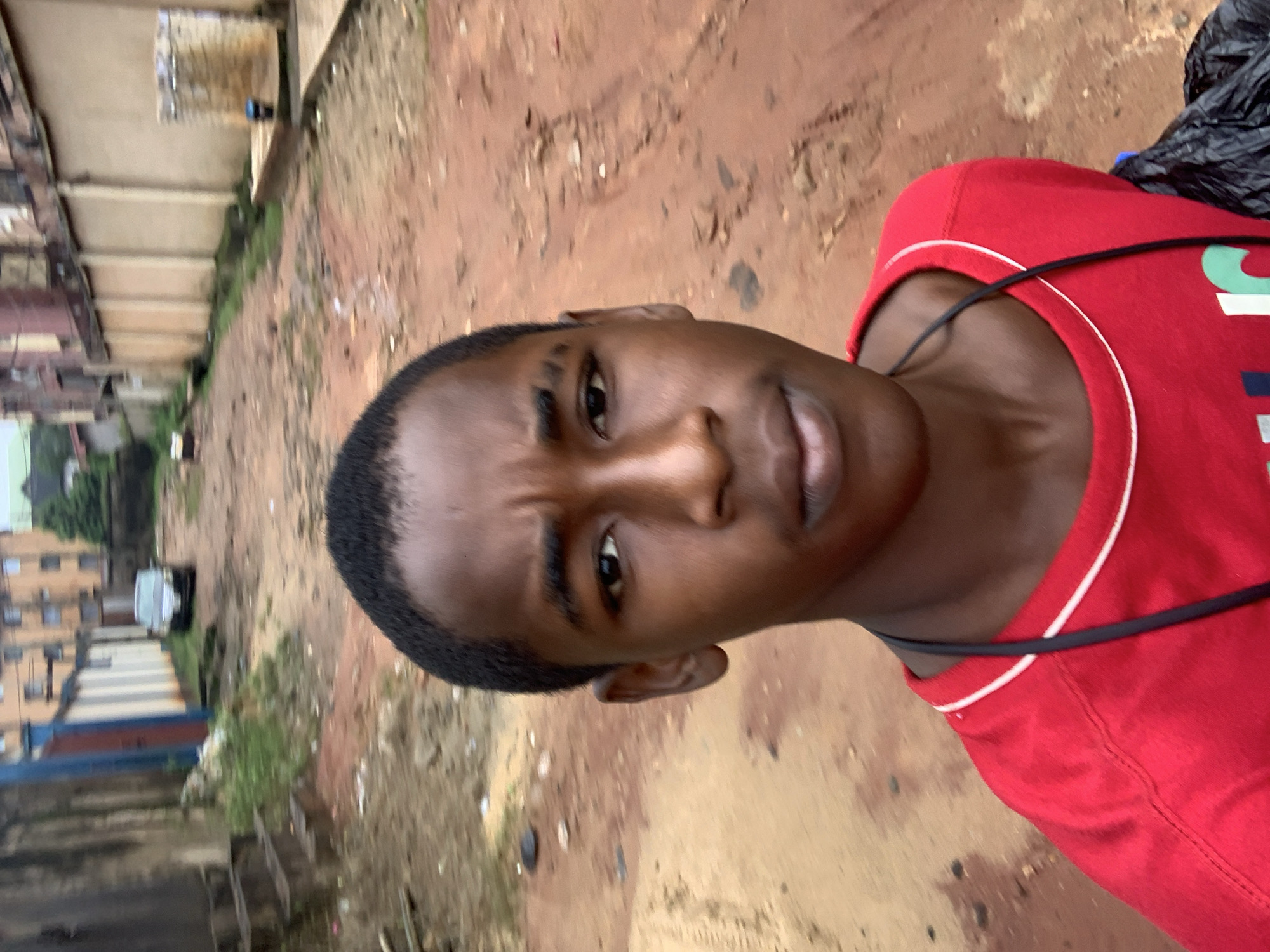 A young Black male, approximately 10-12 years old, is the central subject of this selfie. He is wearing a red t-shirt with a white and black trim around the collar. A black cord or lanyard is visible around his neck. The background suggests an outdoor urban environment in Nkpor, Nigeria, characterized by a reddish-brown dirt road with debris and vegetation. On either side of the road are buildings, some appearing to be residential with multiple stories and balconies, while others are lower concrete structures. A silver car is parked on the left side of the road in the mid-ground, and what appears to be a plastic blue container is visible on the right. The lighting is overcast, suggesting it may be a cloudy day or late afternoon. The overall impression is that of a common street scene in a developing urban area.