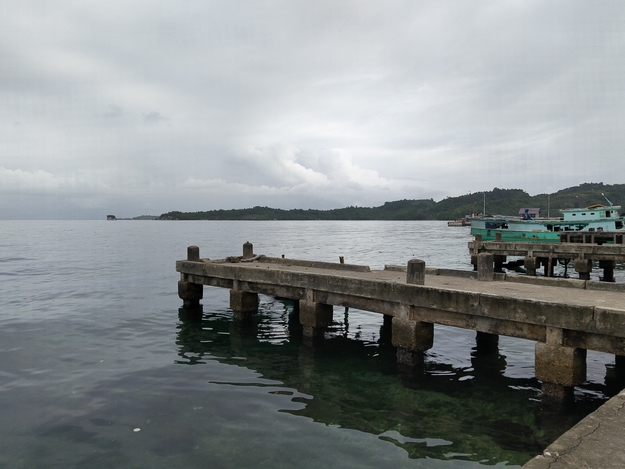 The image shows a concrete pier extending into the water. The pier has a weathered appearance with visible cracks and moss growth on its supports. The water is calm and clear, with the reflections of the pier and the sky visible on its surface. In the background, there is a distant shoreline with lush green vegetation and rolling hills under a cloudy sky. Two boats are docked at another pier in the distance, one of which is light blue and green. The overall scene is one of a peaceful, somewhat overcast day at a coastal location. This scene is typical of the Banggai Islands, known for their natural beauty and coastal infrastructure that supports local fishing and transportation.