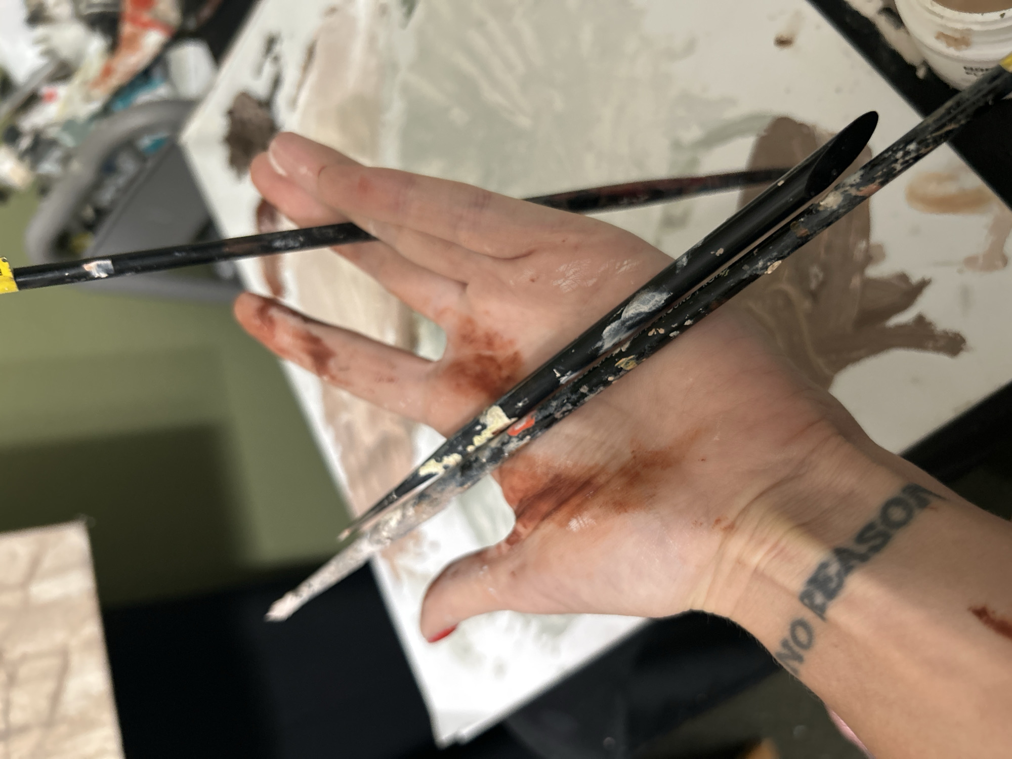 The image shows a close-up of a person's hand holding two paintbrushes. Brown paint smears are visible on the hand. The person has a tattoo on their wrist that says "NO REASON". The paintbrushes are black and covered in paint. In the background, there is a paint palette with various shades of brown paint. There is no indication of a specific location, and there is no information to confirm the location as Cape Coral, United States of America.