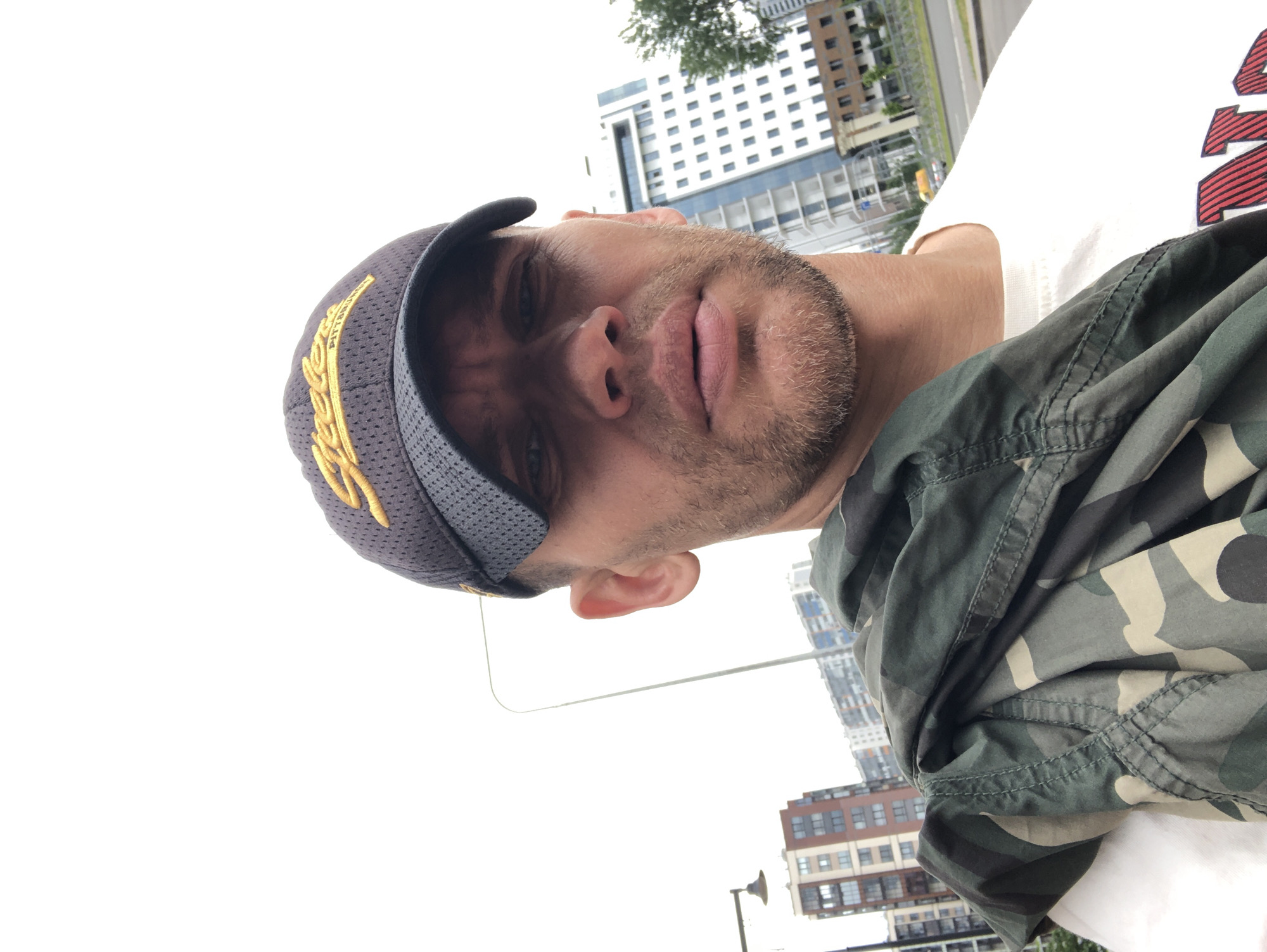 A man wearing a black and yellow baseball cap and a camouflage jacket over a white t-shirt poses for a selfie outdoors. He has short brown hair, stubble, and fair skin. In the background, modern buildings and a street are visible, suggesting an urban environment. The lighting is overcast. The image appears to have been taken in Saint Petersburg, Russia.