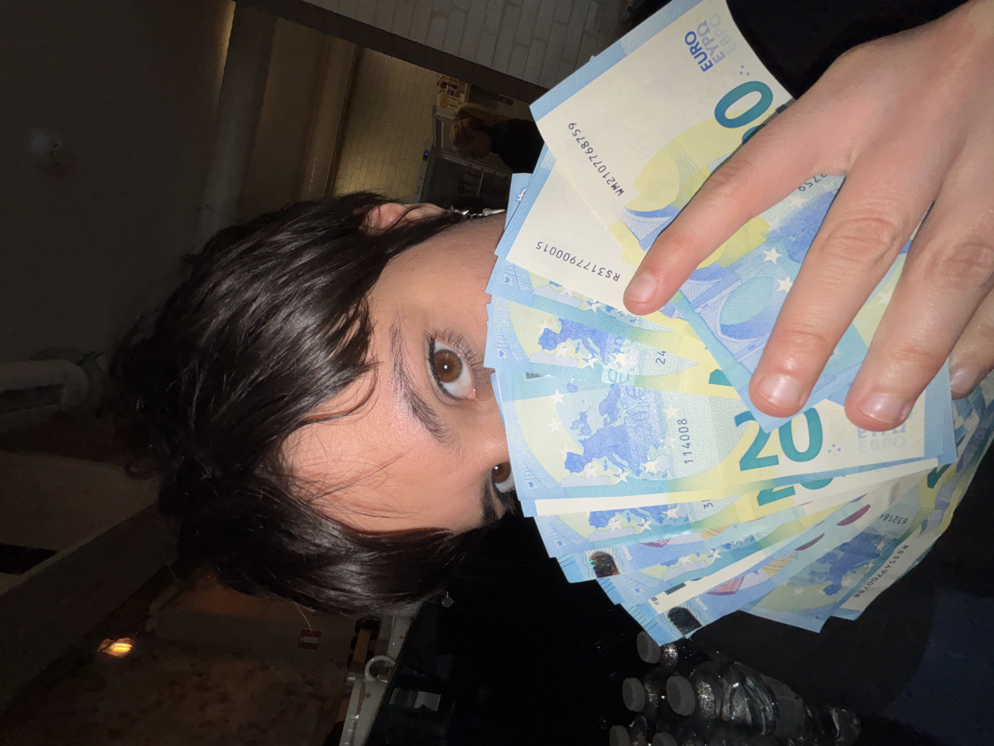 A person with short dark hair and brown eyes is holding a stack of 20 Euro banknotes in their right hand, partially obscuring their face. The banknotes appear to be new. The person is wearing a dark top. The background is dimly lit, with a glimpse of what appears to be a bar or restaurant interior with other people visible. The image is taken from a low angle, tilted slightly. The presence of Euro banknotes suggests a location where this currency is accepted. Considering the user's request for a location in Sofia, Bulgaria, it is plausible the image was taken in a bar, restaurant, or business establishment in Sofia that accepts Euro currency.