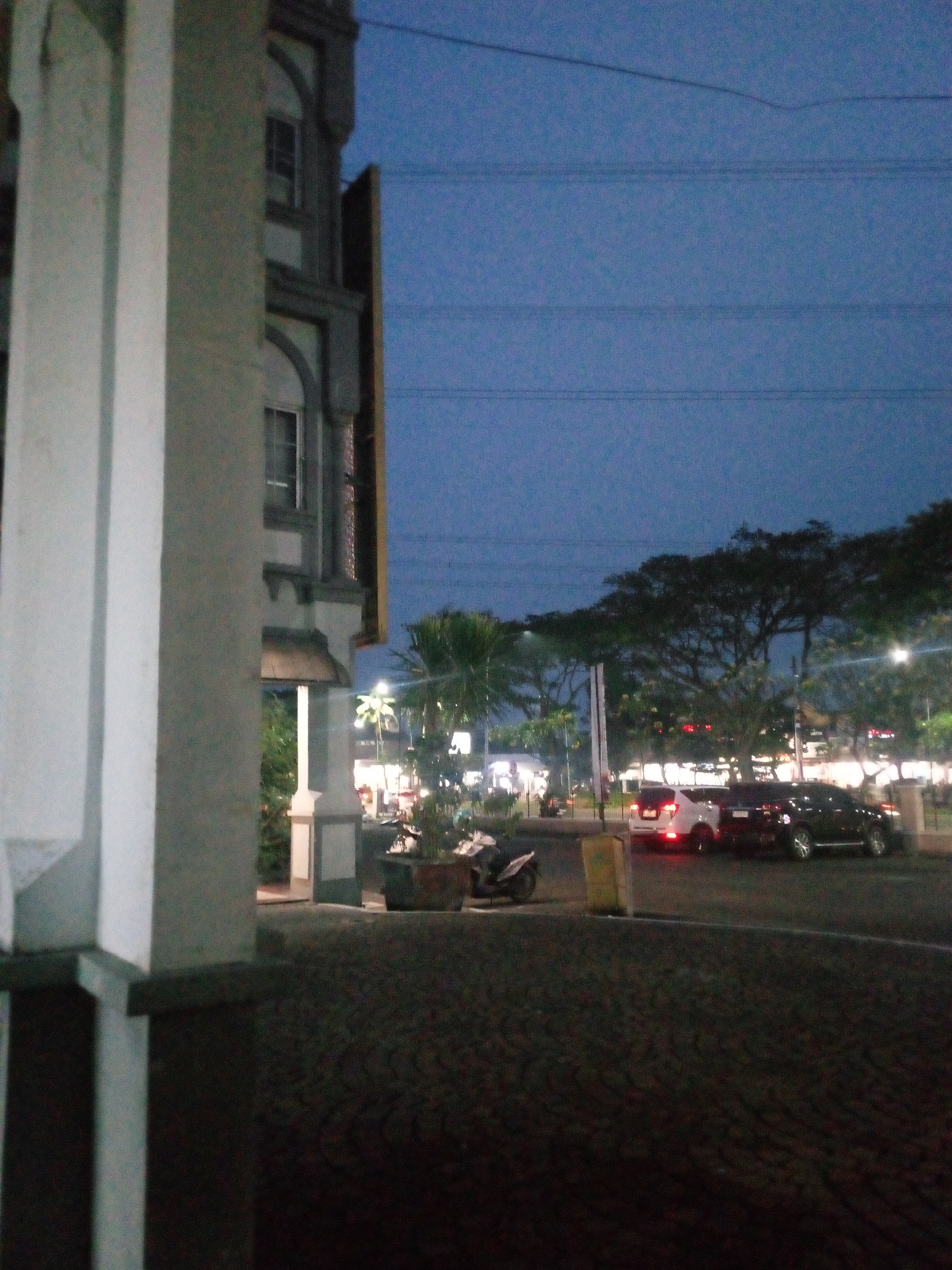 The image depicts an outdoor scene at dusk or night in Cikupa, Indonesia. On the left, a portion of a building with a light-colored column in the foreground is visible. The building has architectural details with arched windows. To the right of the building, a street scene unfolds. Several vehicles are present, including a white SUV and a black SUV, parked or driving on the road. A motorcycle is also parked on the side. Trees and streetlights illuminate the area, casting a warm glow on the surroundings. The sky is a deep blue, indicating twilight or nightfall, with visible power lines stretching across it. The ground appears to be paved, possibly with cobblestones or patterned asphalt. The overall atmosphere suggests a commercial or residential area with some activity.