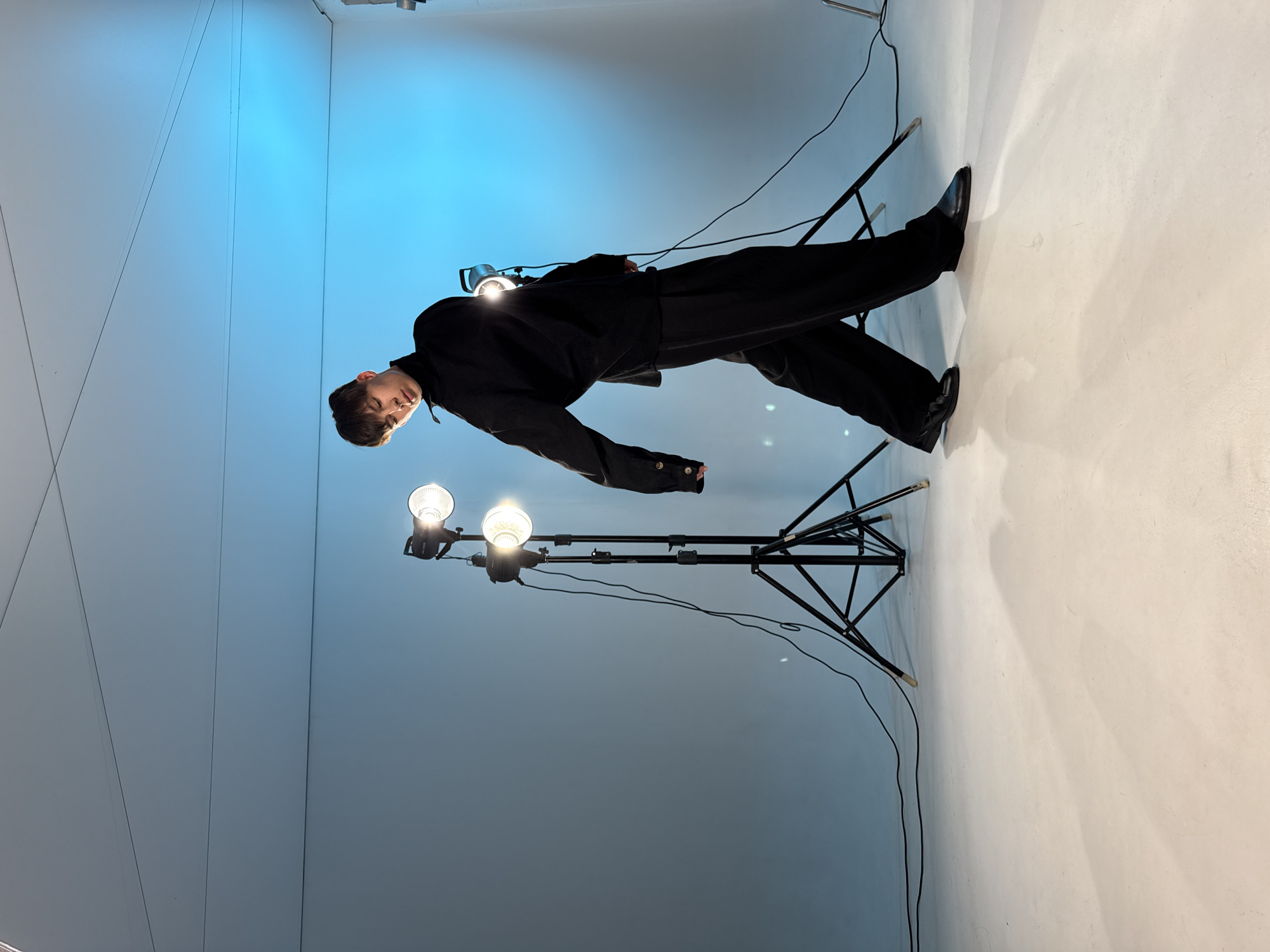 A man in a black suit and shoes is lying on a chair in a photography studio. Two light stands with lamps are positioned near him. The backdrop of the studio transitions from a light blue to a white gradient. The location is not specified as Chaikovsky, Russia, but assumed to be a photography studio setting.