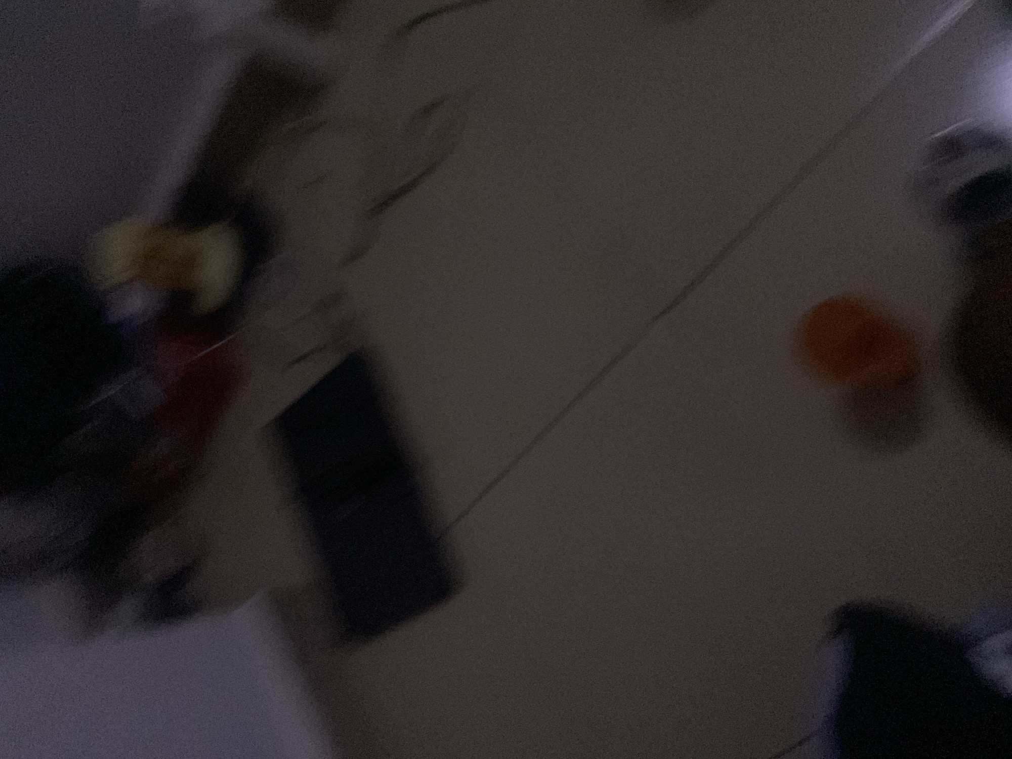 The image depicts an indoor scene, possibly a bedroom or living area, with a floor and some scattered objects. The lighting is dim, and the image is blurry, suggesting movement during capture or low light conditions.
In the center of the frame, there is a dark, rectangular object that appears to be a laptop, lying open on the floor. A thin cable extends from the laptop diagonally across the floor.
To the lower right of the laptop, there is a small, round, orange object, possibly a ball or a piece of fruit.
In the upper right portion of the image, there is a blurred yellowish object, its nature unclear due to the blur.
The floor appears to be tiled or made of a smooth material, with a light brown or beige color. The walls are not clearly visible, but there is a hint of a lighter colored surface on the left side of the image, suggesting a wall or furniture.
There are no discernible people or clear interactions in the scene. The overall impression is that of a disheveled or temporarily occupied space. There are no identifiable elements that would allow for the determination of its location within Howayi, Nigeria.