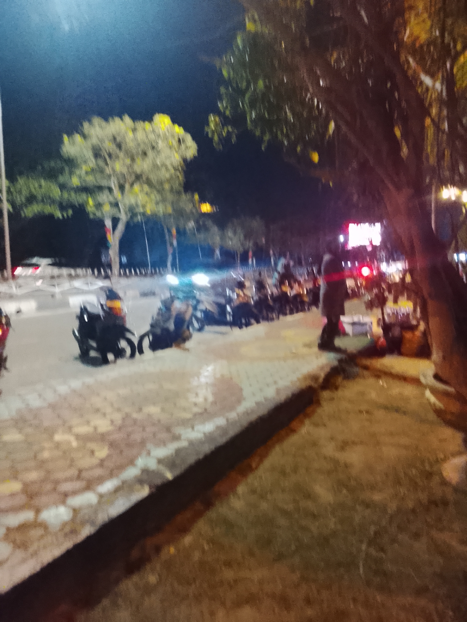 The image depicts a nighttime street scene in Teluk Air, Indonesia, characterized by its blurry, low-light conditions. In the foreground, a paved walkway with patterned bricks leads towards the street, where several motorcycles are parked in a row. Beyond the motorcycles, a person in dark clothing with a red sash is visible standing near a bright red light. The background features trees with illuminated foliage and distant city lights, suggesting an urban or semi-urban environment. The overall impression is that of a casual gathering or a roadside market at night.